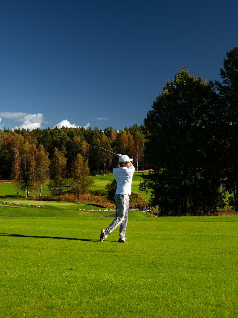 Golfer in white shirt and cap swings club on green fairway, with autumn trees and deep blue sky.