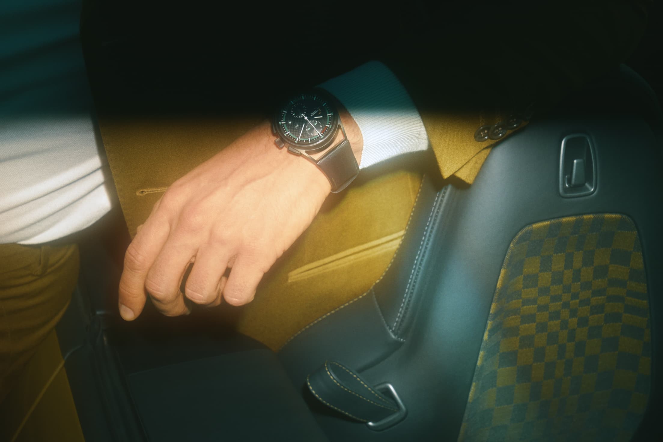 Close-up of a man wearing an olive-colored suit and sitting in a Porsche, wearing a Porsche Design watch
