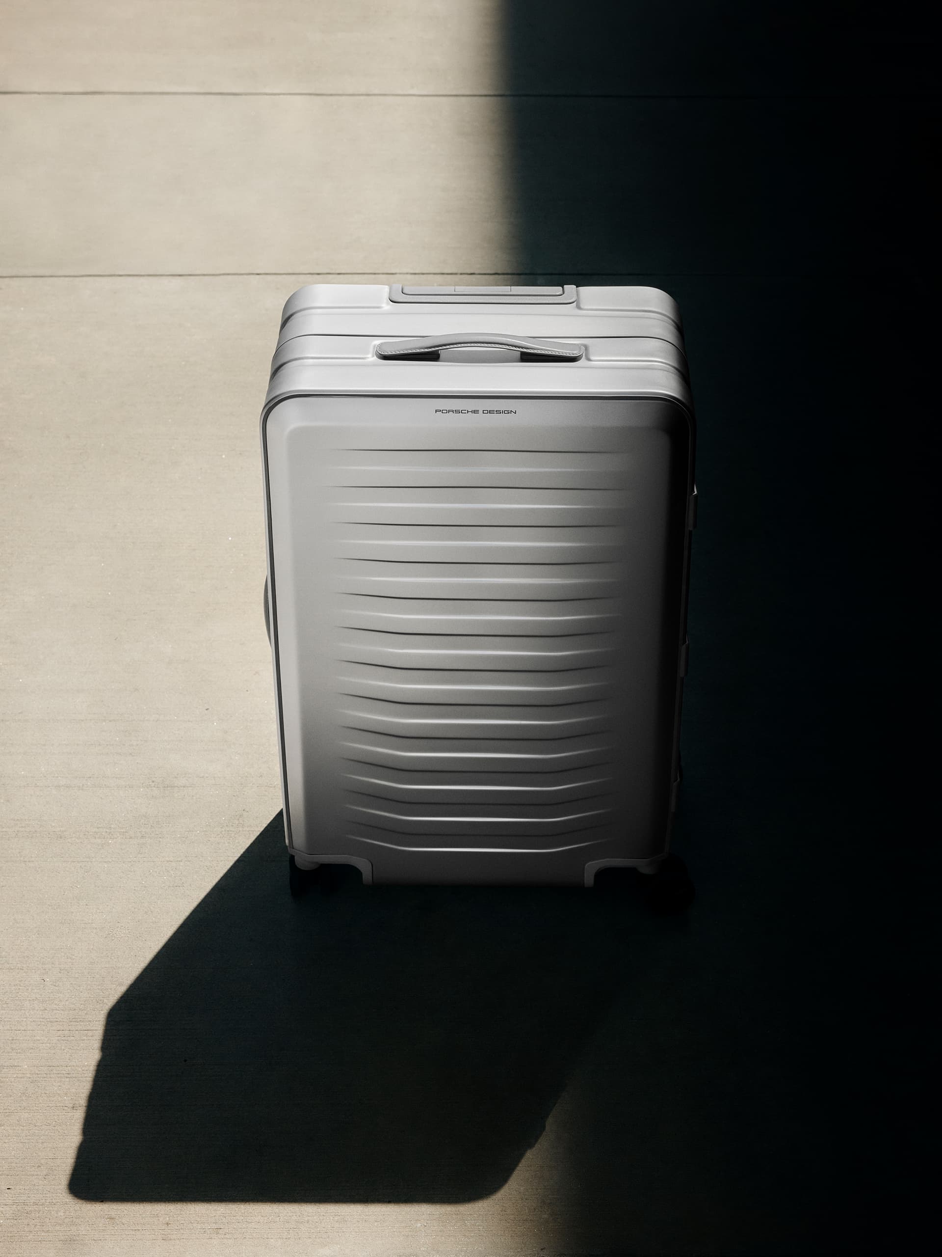 Silver hard-shell suitcase with ribbed texture and carry handle, in a cone of light on a concrete floor.