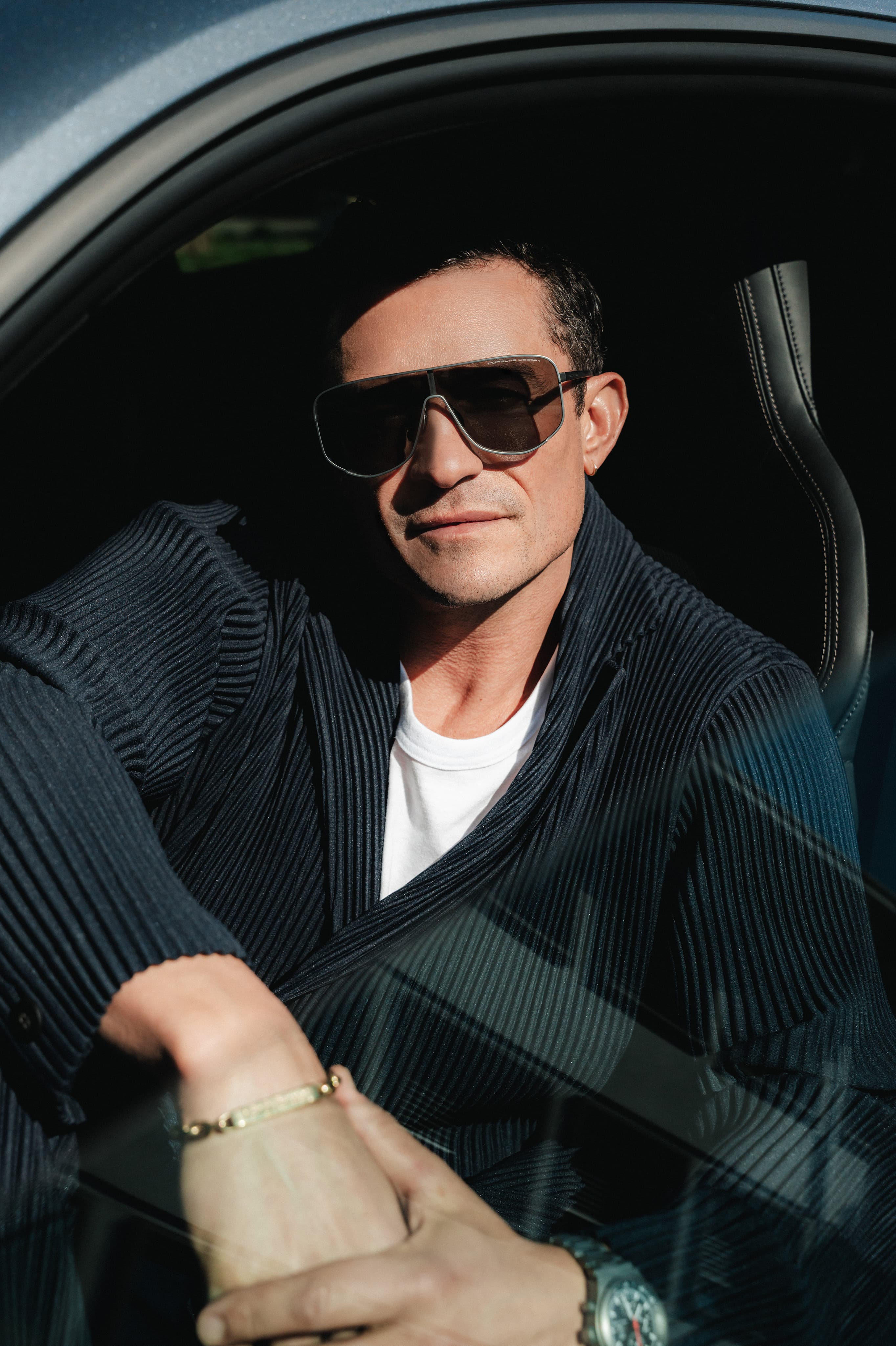 Orlando Bloom im Auto, trägt große, eckige Porsche Design Sonnenbrille mit dunklen Gläsern, schwarzen Cardigan und weißes T-Shirt.