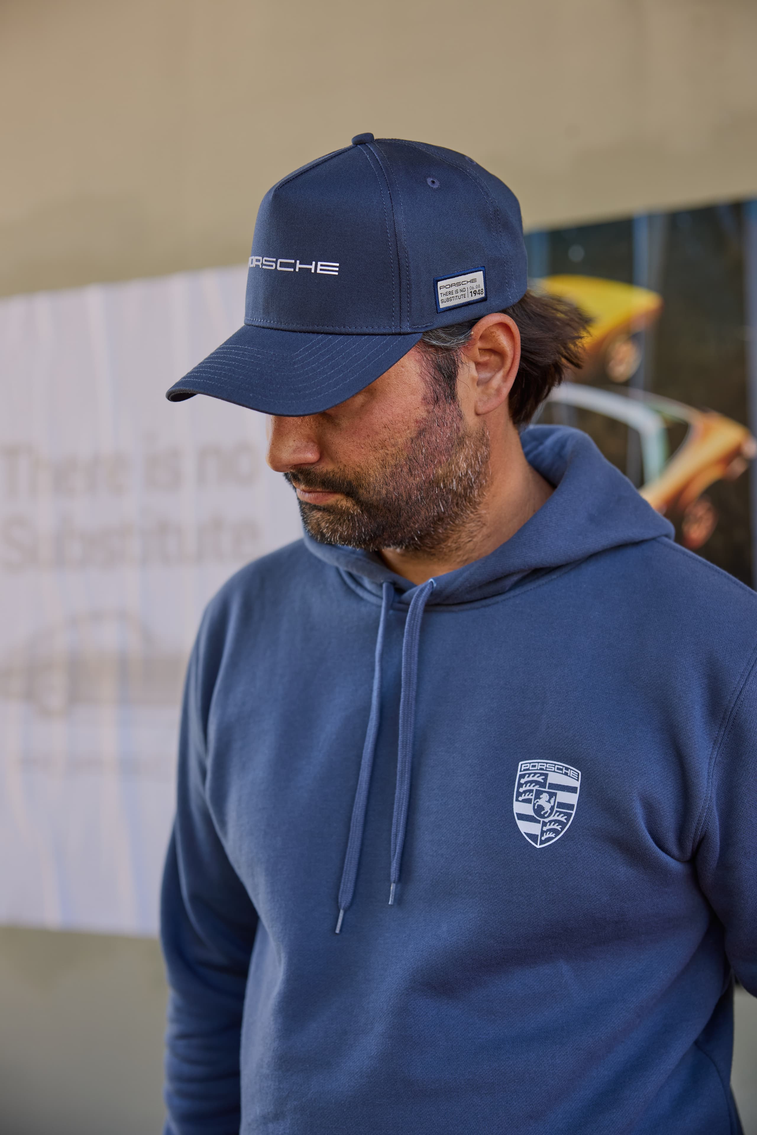 Man wearing a dark blue Porsche cap with a patch and a dark blue hoodie with the Porsche crest, side view.