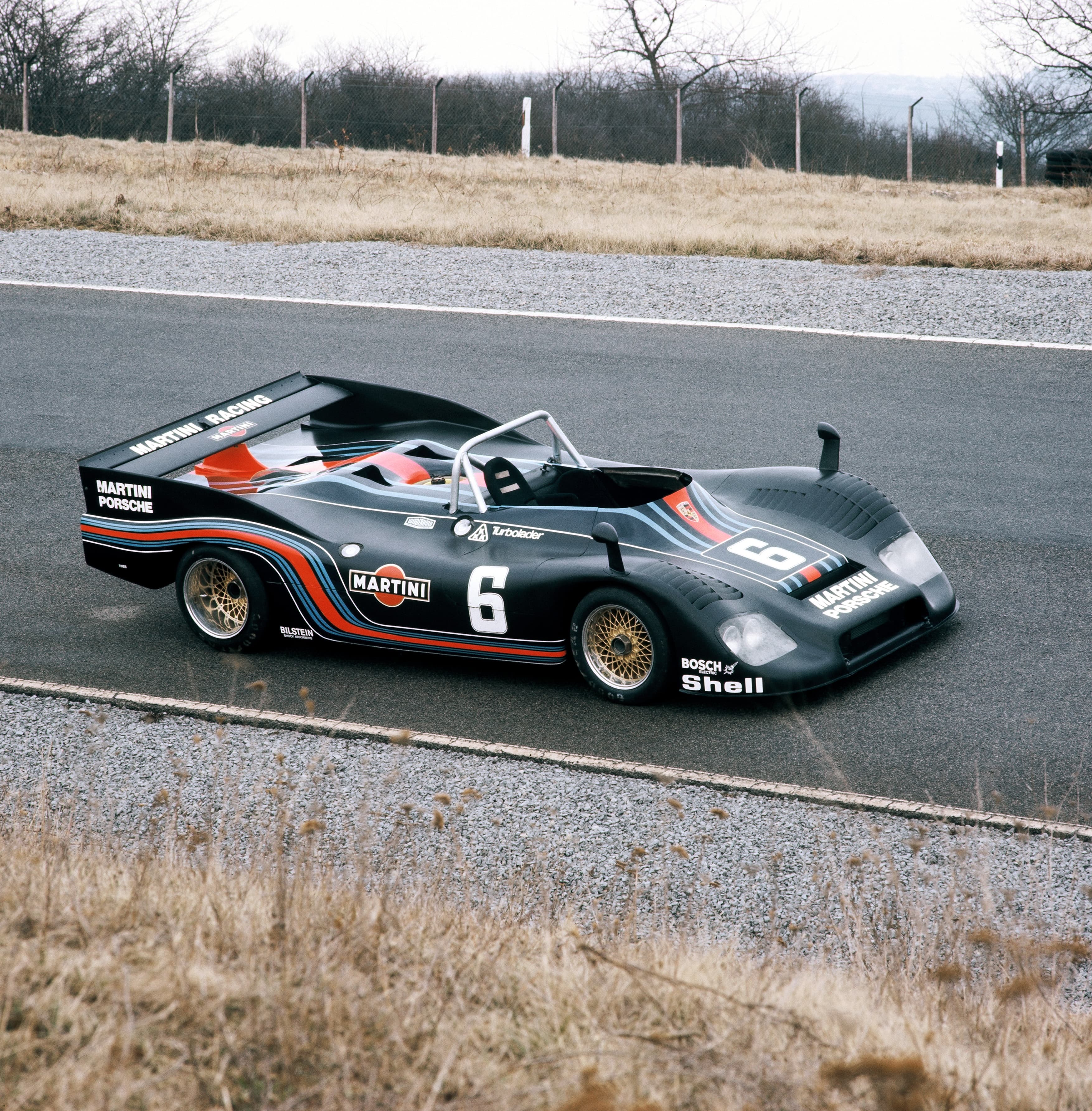 Vintage-Porsche-Rennwagen mit Martini-Lackierung, Nummer 6, auf einer Strecke mit trockenem Gras und kahlen Bäumen im Hintergrund.