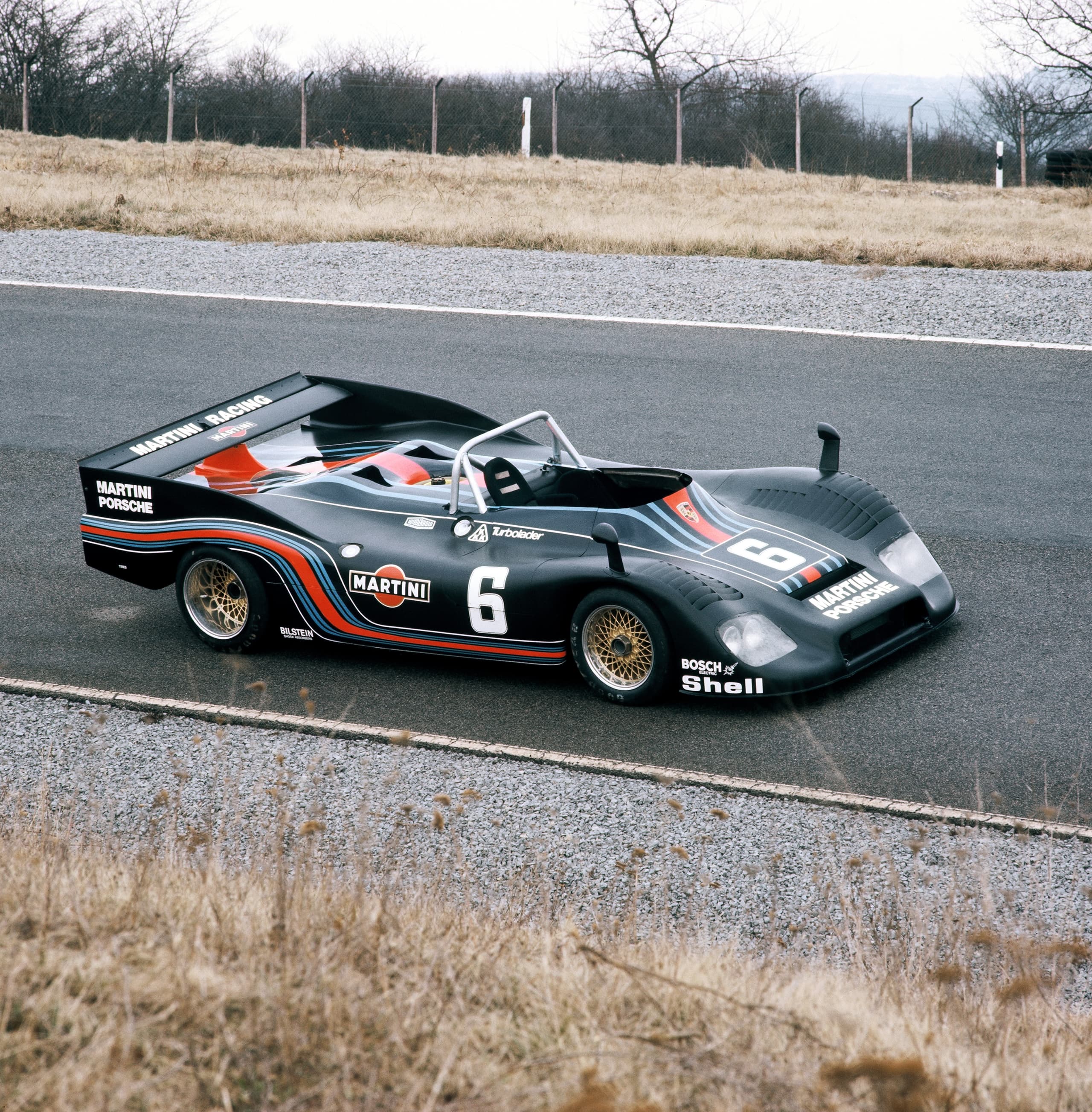 Vintage Porsche race car with Martini livery, number 6, on a track with dry grass and bare trees in the background.