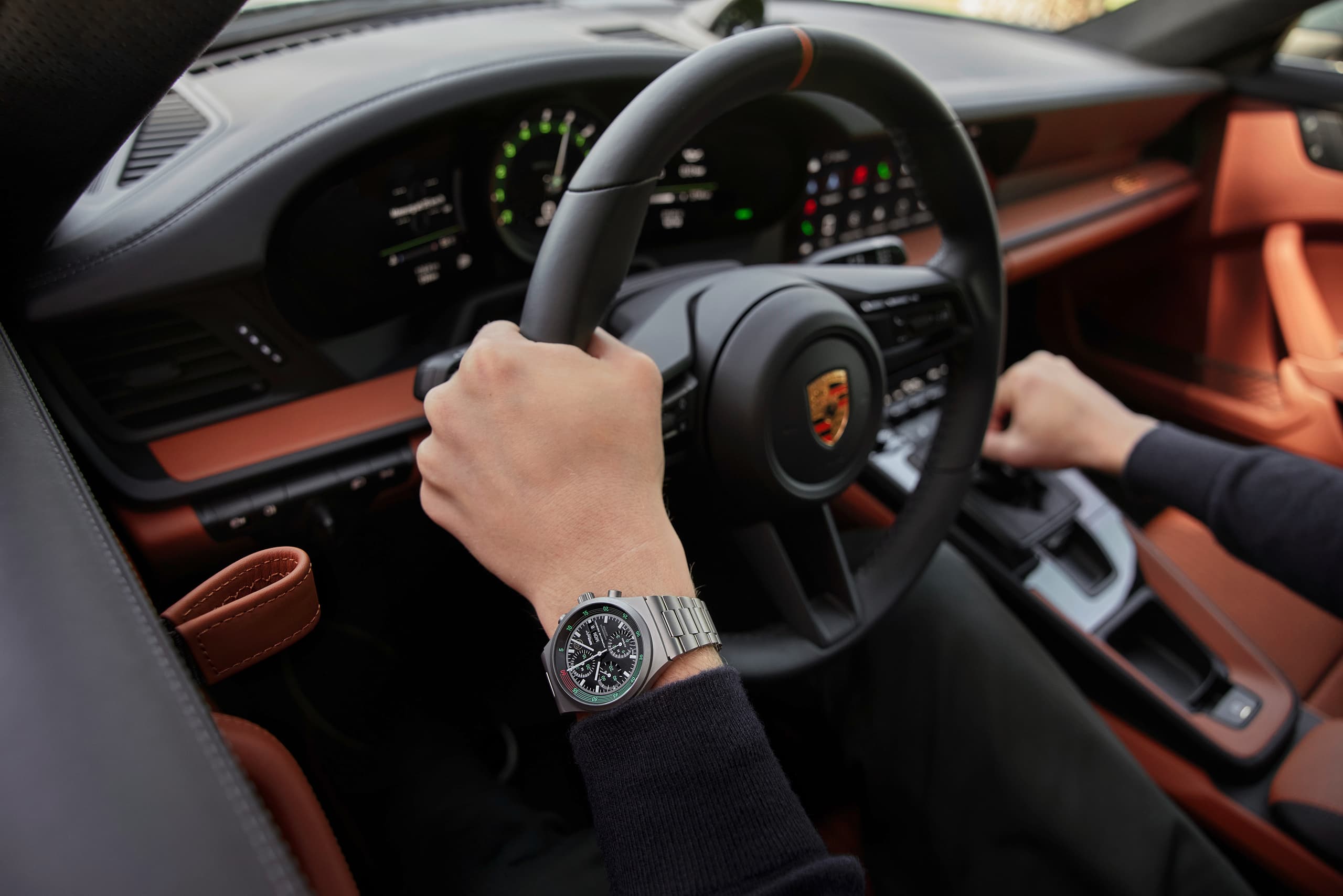 Dłonie na kierownicy Porsche, na nadgarstku chronograf ze stali nierdzewnej, wnętrze w czarnej i koniakowej skórze.