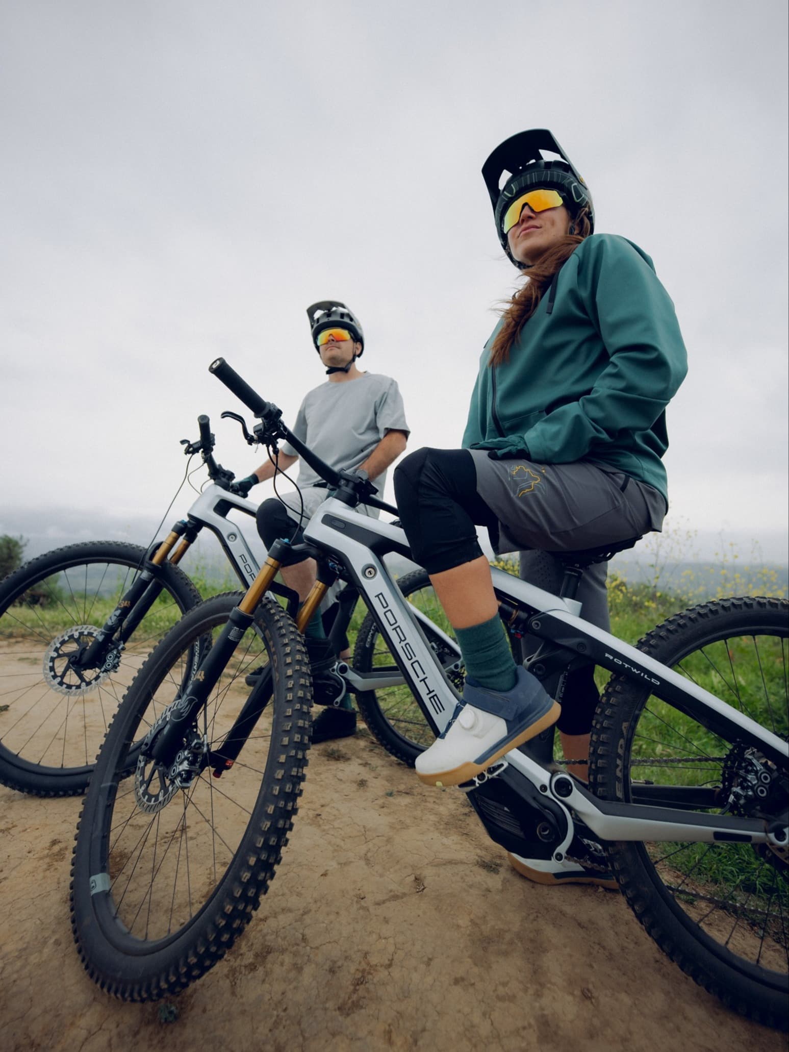 Due mountain biker con casco e occhiali a specchio su e-bike grigie, in pausa su un sentiero sterrato sotto un cielo grigio.