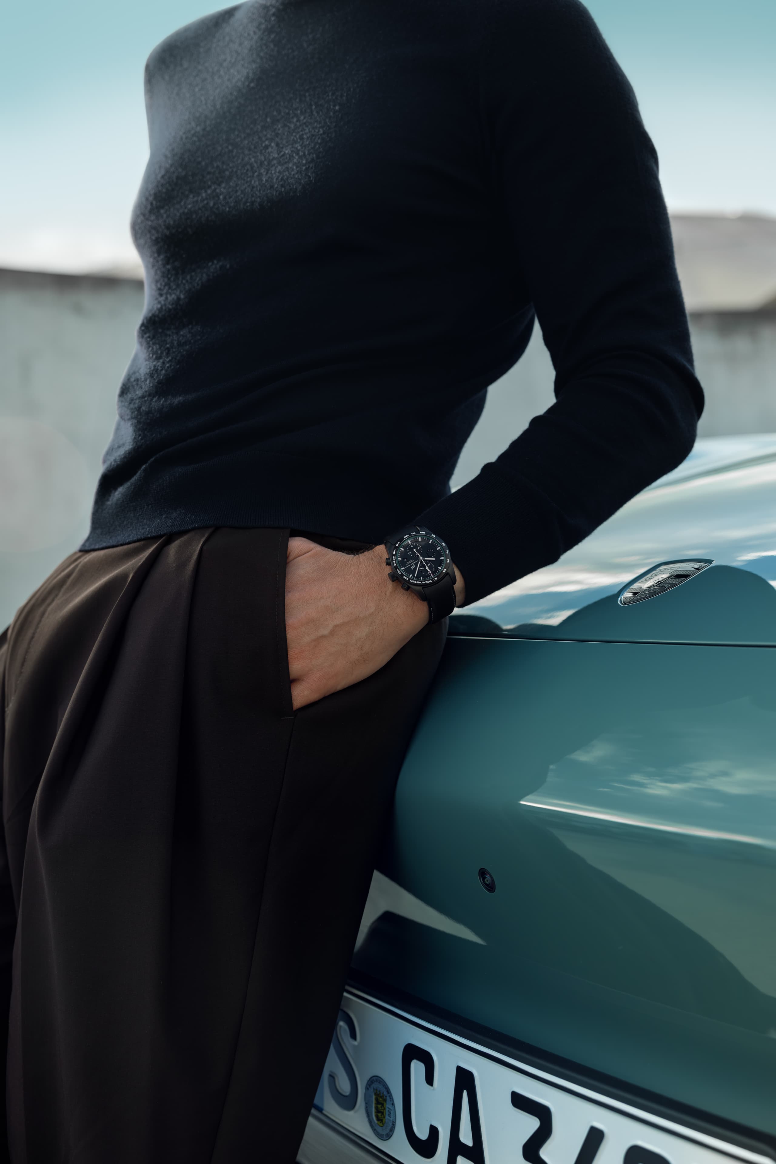 Image shows man wearing a blue custom-build Porsche Design Timepieces colormatching to the Porsche Cayennne next to him (blue)