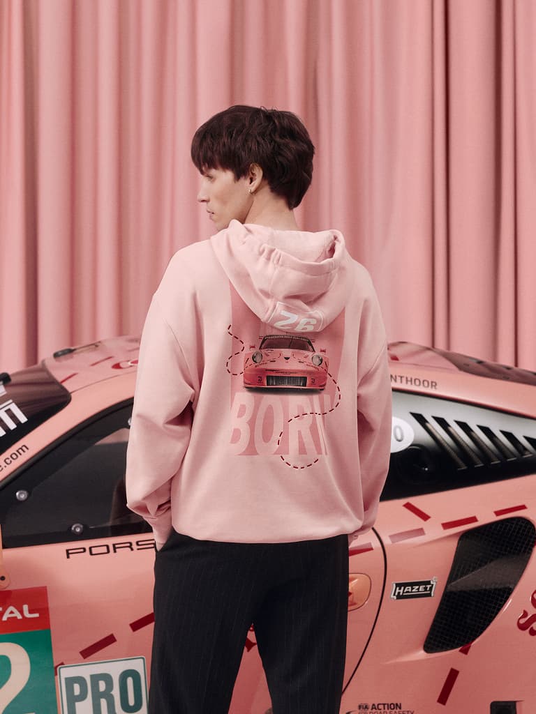Pessoa com hoodie rosa com gráfico de carro ao lado de carro de corrida Porsche rosa, cortina rosa ao fundo.