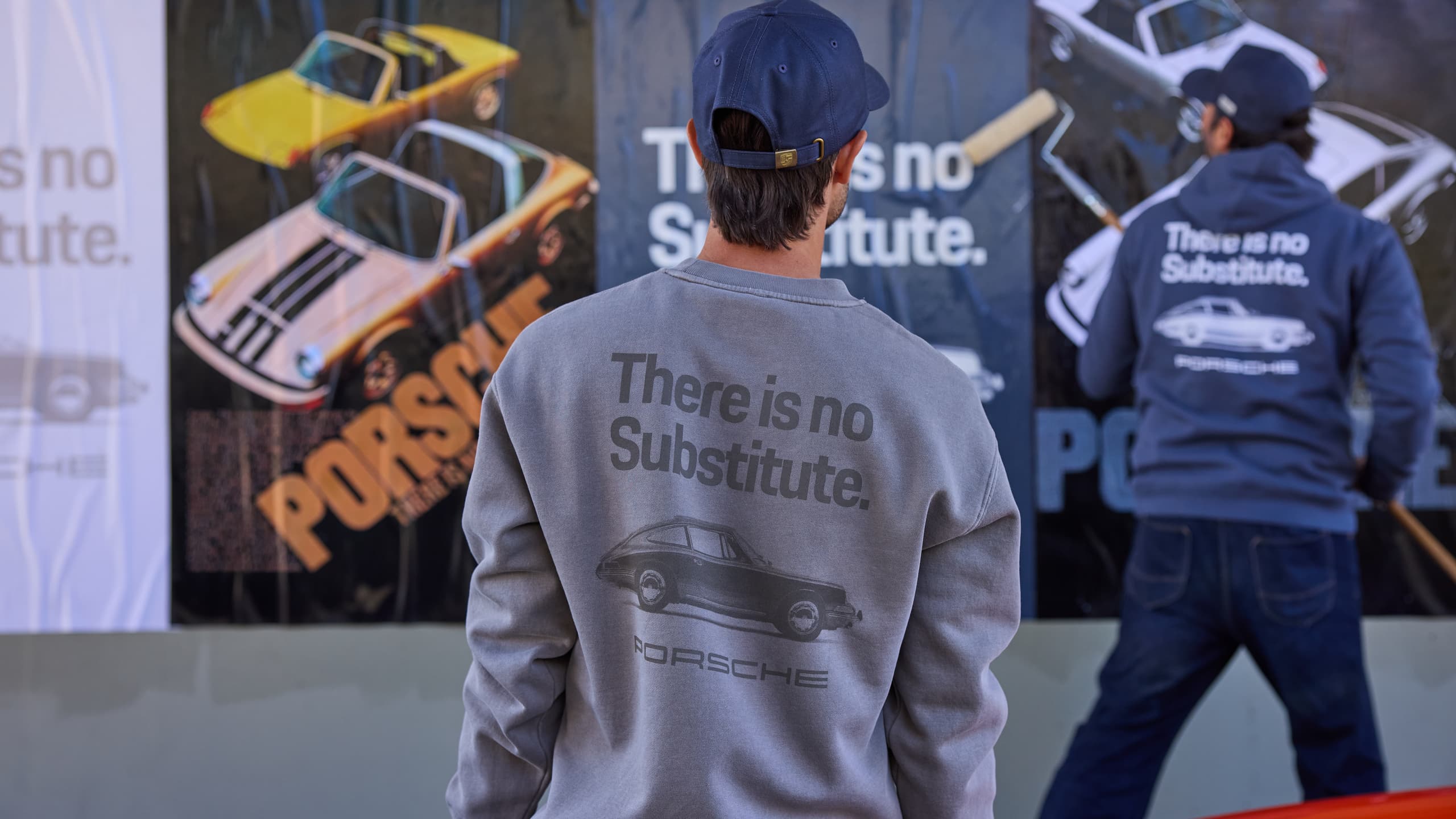 Man in a gray Porsche sweatshirt and blue cap in front of a poster wall featuring sports car motifs, with a second person on the right.