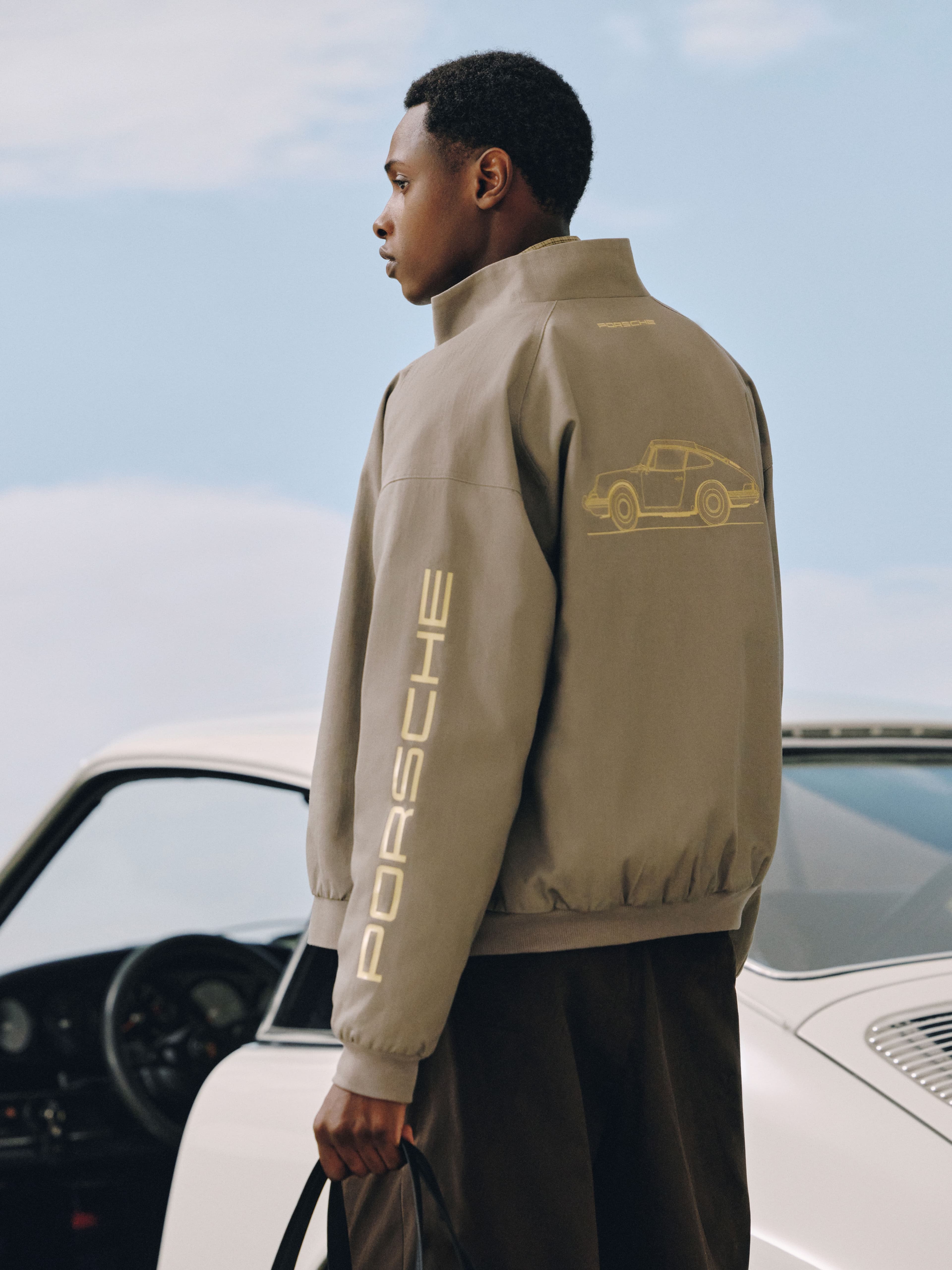 "Man in a beige Porsche jacket with a back print, standing sideways next to a white sports car under a blue sky."