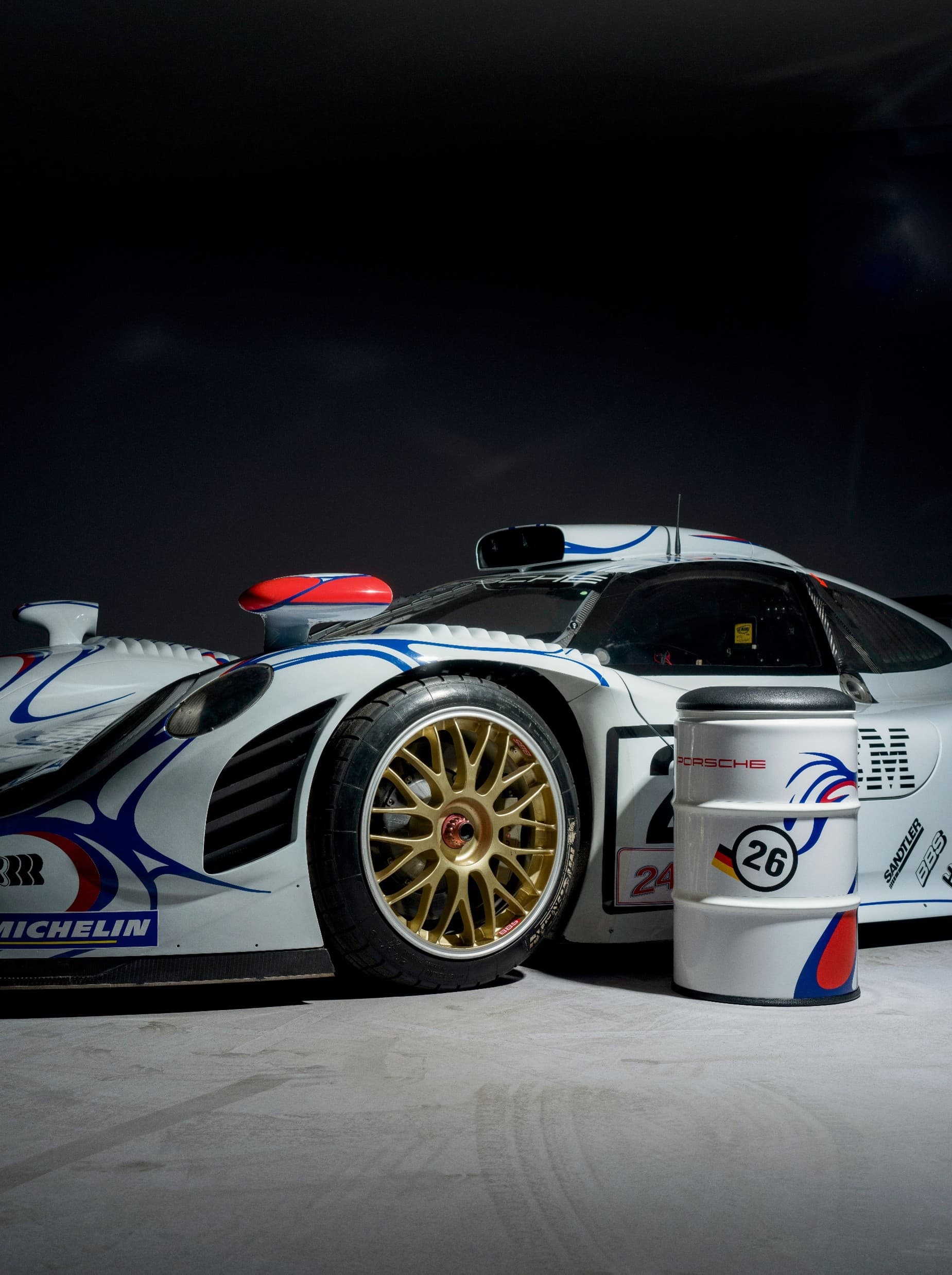 Carro de corrida Porsche branco com rodas douradas, detalhes em vermelho e azul, ao lado de um barril correspondente com o número 26 em fundo escuro.