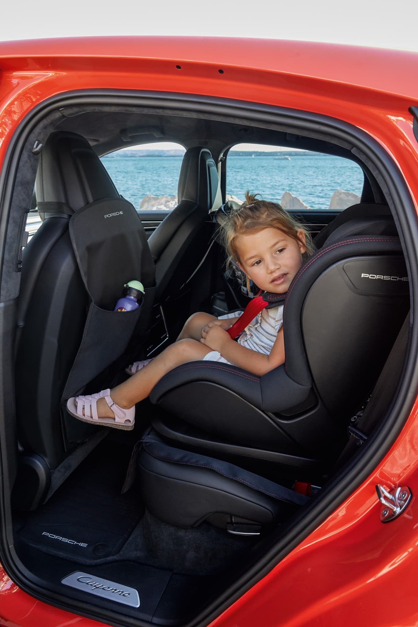 Child in a black Porsche child seat in the rear of a red Porsche Cayenne, looking outside, sea in the background.
