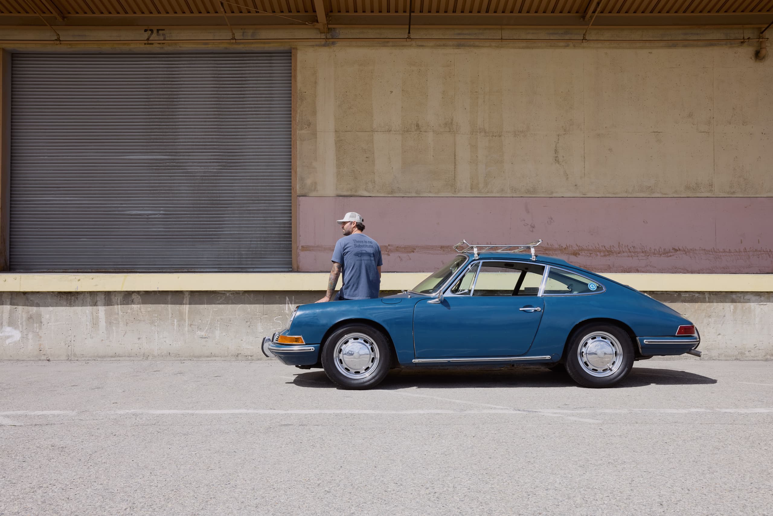 Man in a blue T-shirt next to a blue classic Porsche 911 with a roof rack in front of an industrial building.