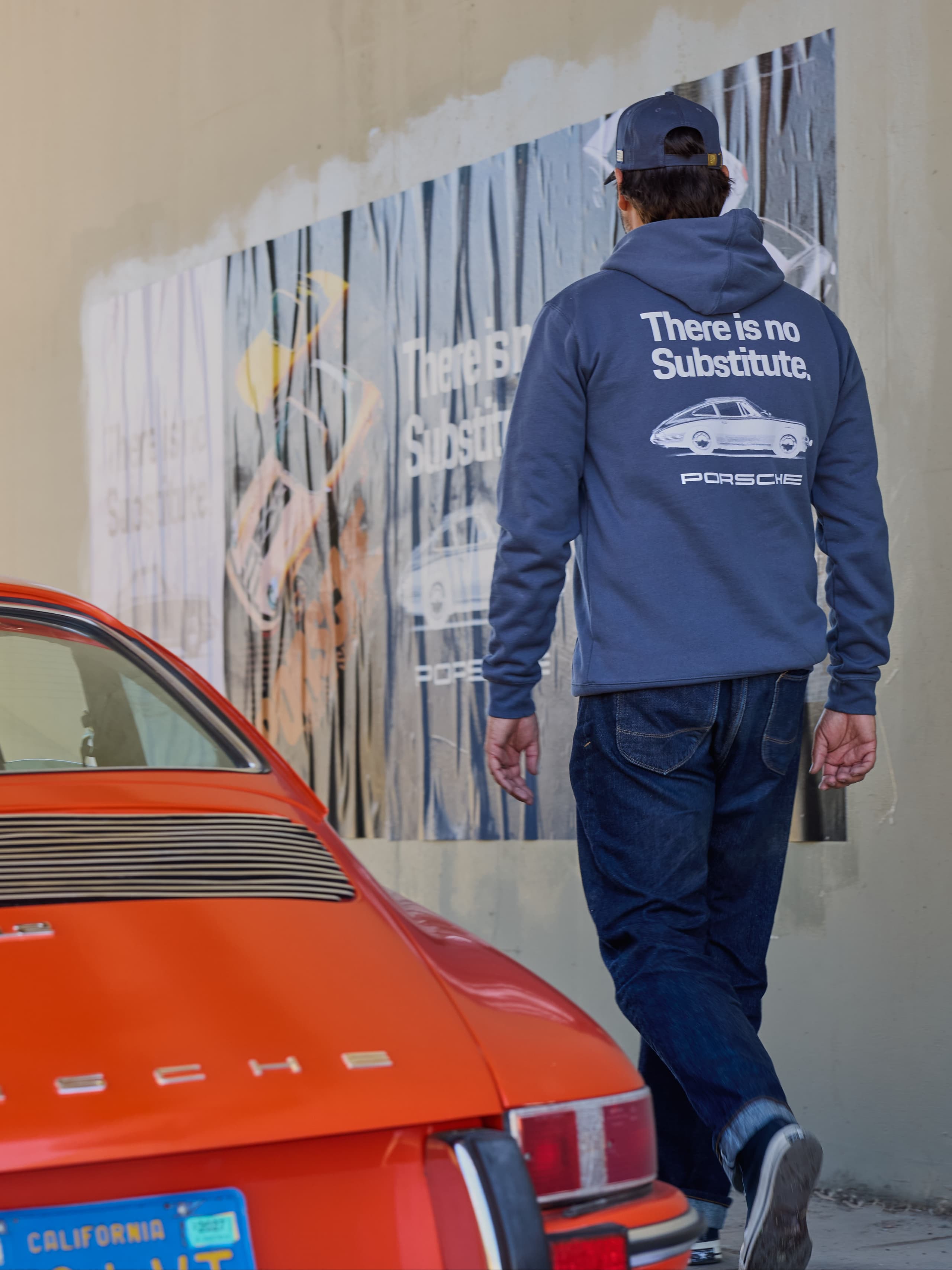 Man in a navy Porsche hoodie and cap in front of a wall poster, next to the rear view of an orange Porsche 911.