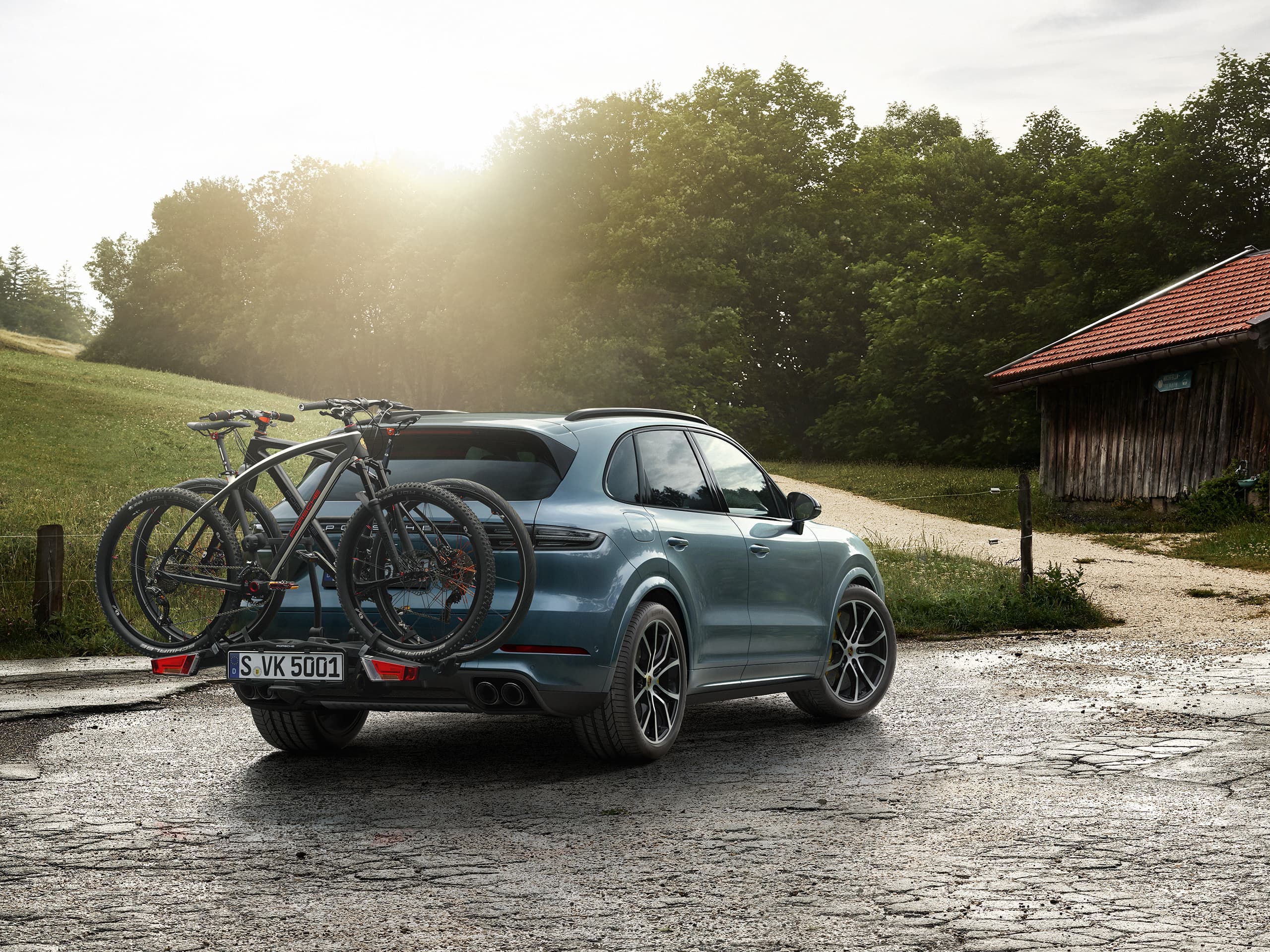 Blue Porsche SUV with two mountain bikes on a rear bike rack, parked on a country road at sunset.