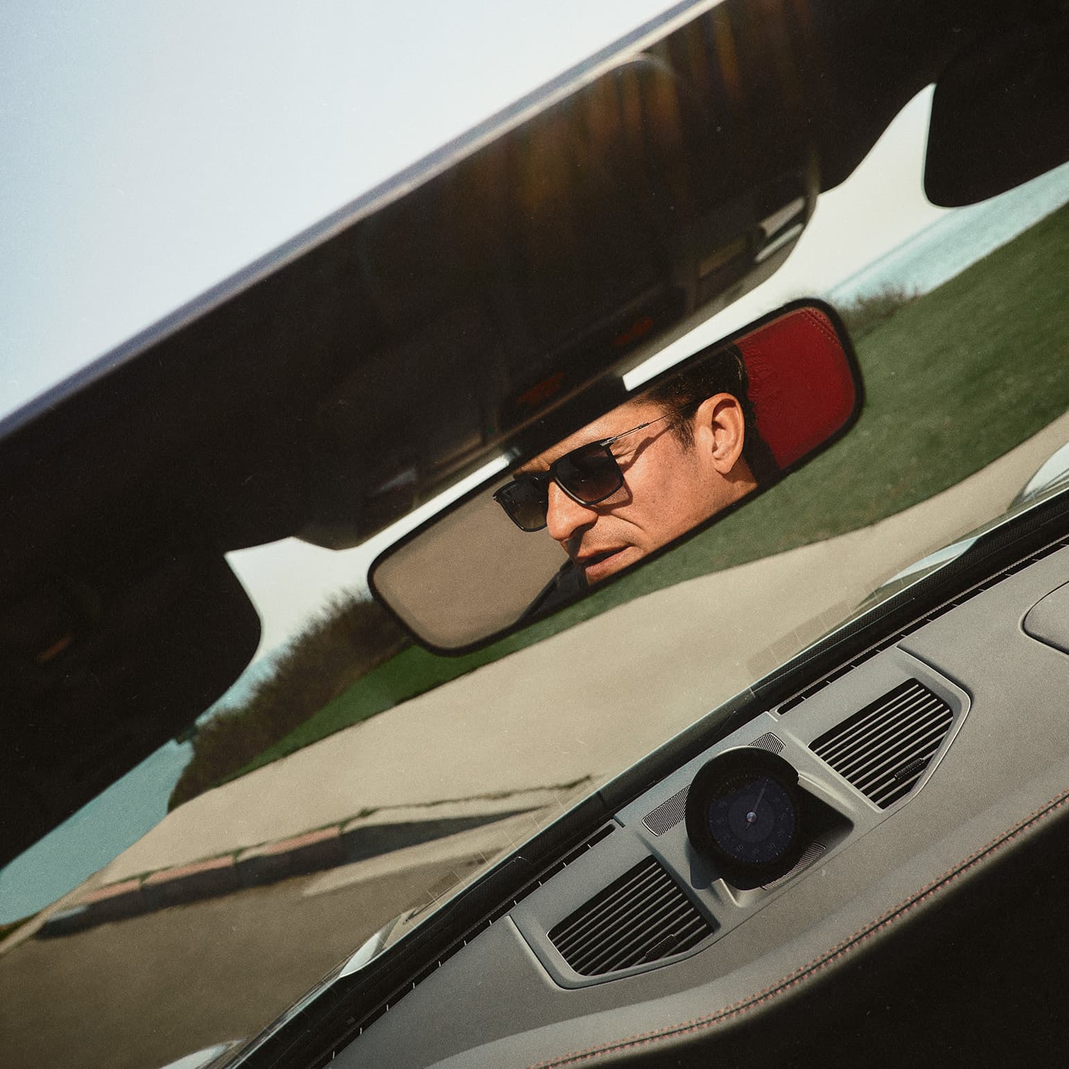 Orlando Bloom sits in a Porsche convertible and wears Porsche Design sunglasses. You can see this through the rear-view mirror.