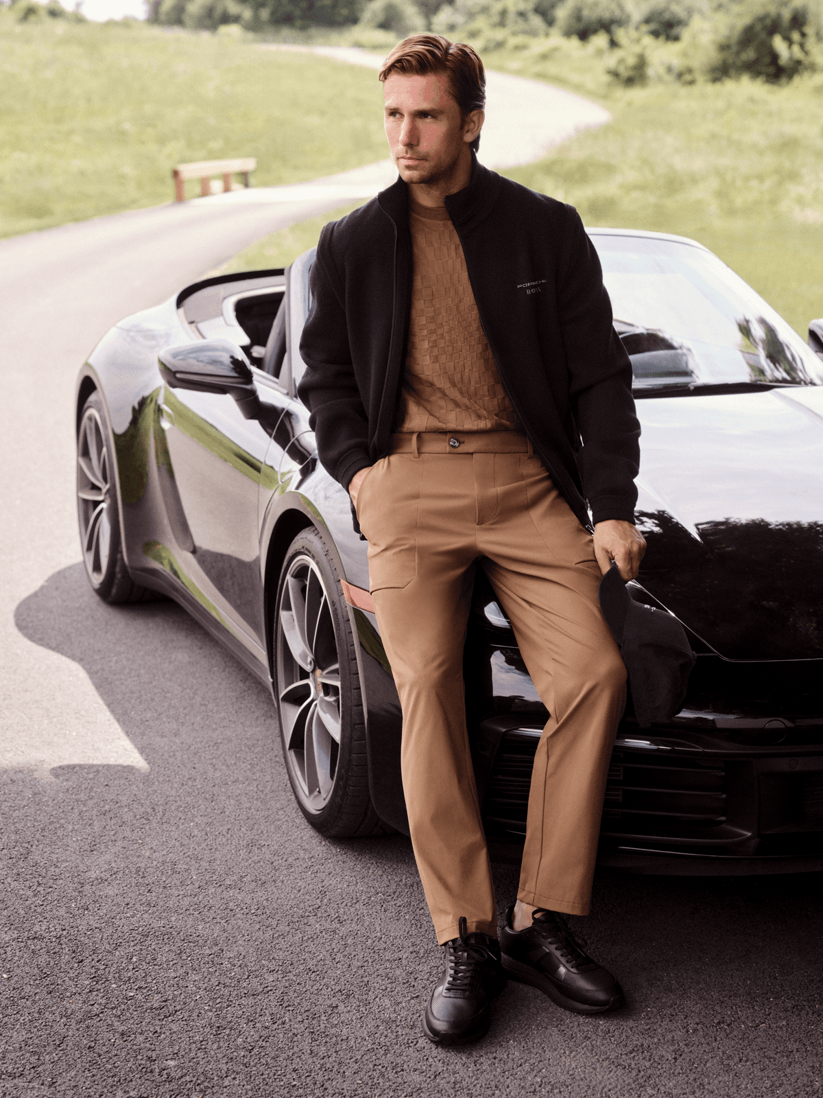 Man in a black jacket and beige outfit leaning against a black Porsche convertible on a winding country road.