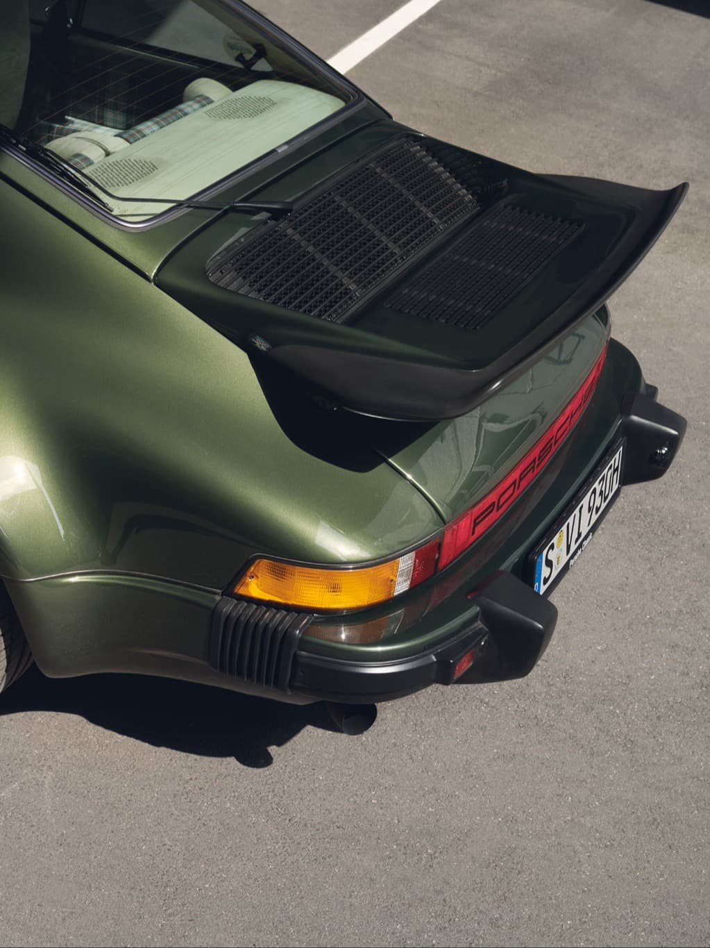 Bakifrån sedd olivgrön Porsche 911 med svart bakspoiler och röd ljuslist, på asfalt.