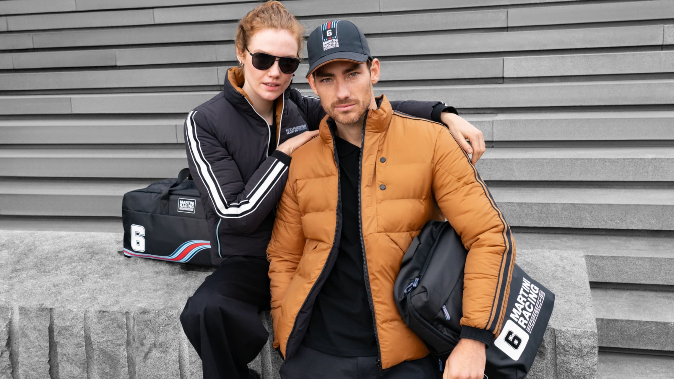 Two people in Porsche jackets and caps, sitting on steps with Martini Racing bags, against a gray wall.