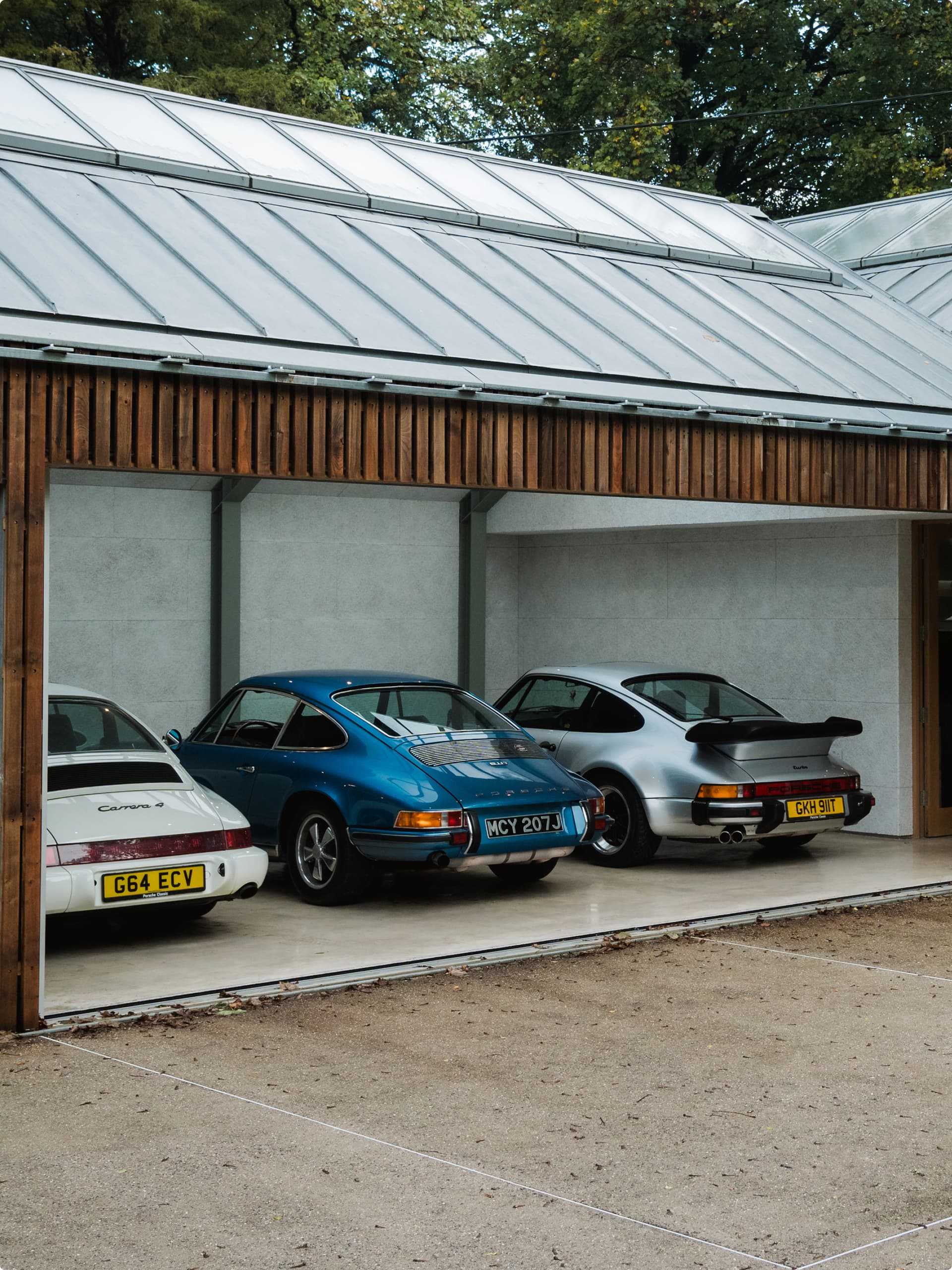 Drei klassische Porsche-Autos in Weiß, Blau und Silber, geparkt in einer modernen Garage mit Holzfassade und Glasdach.