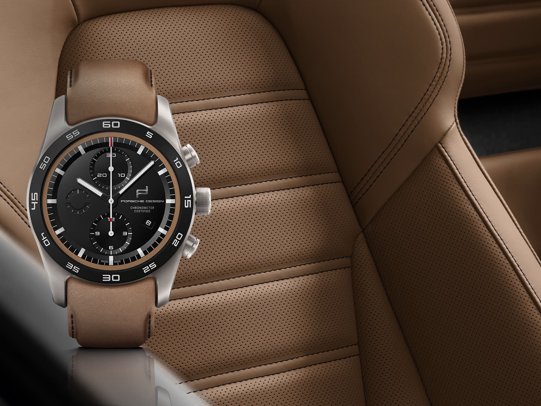 Chronograph watch with black dial and brown leather strap, shown in front of a tan perforated leather car seat.