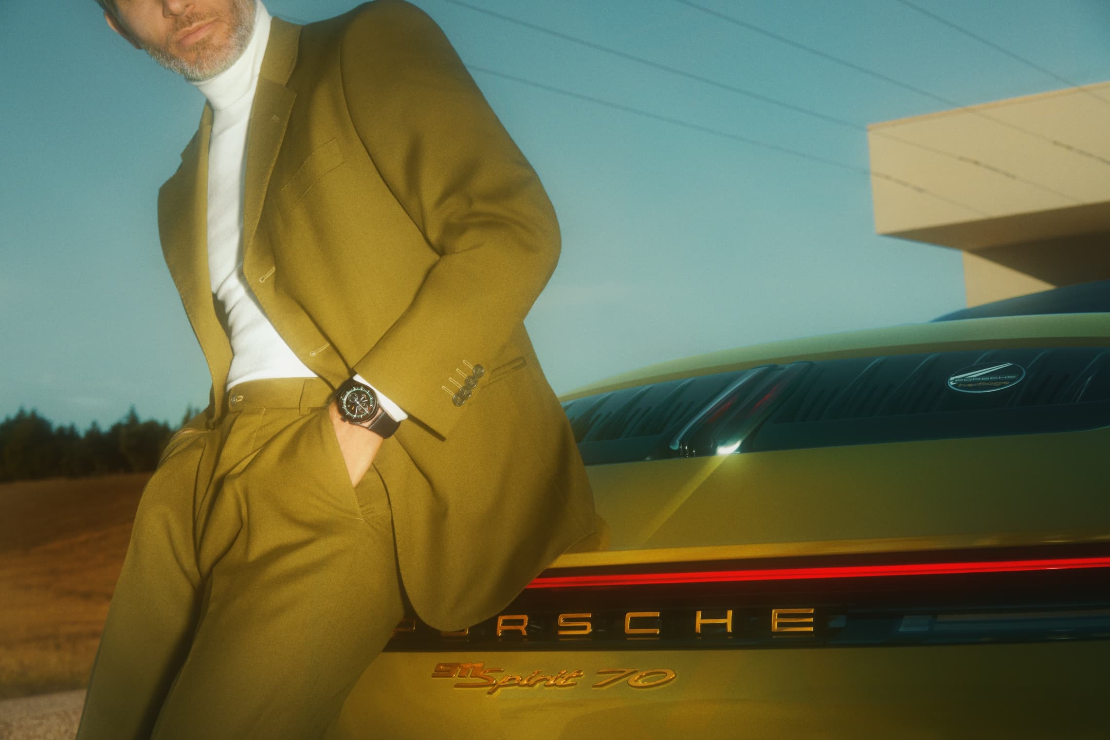 Man wearing an olive-colored suit and leaning against a Porsche, wearing a Porsche Design watch
