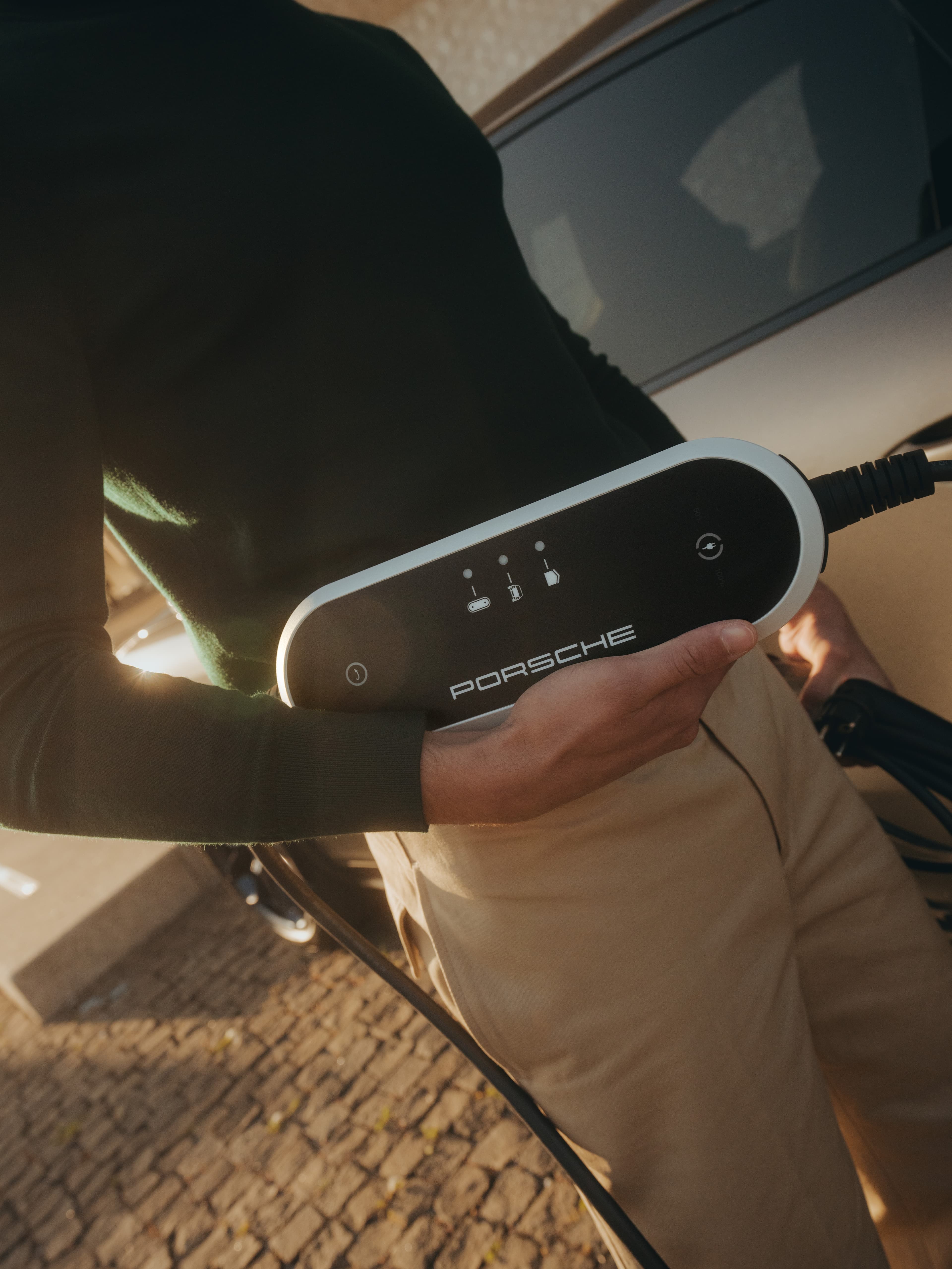 Person holding a Porsche charging cable, wearing a dark sweater and beige pants, standing by a car on a cobblestone street.