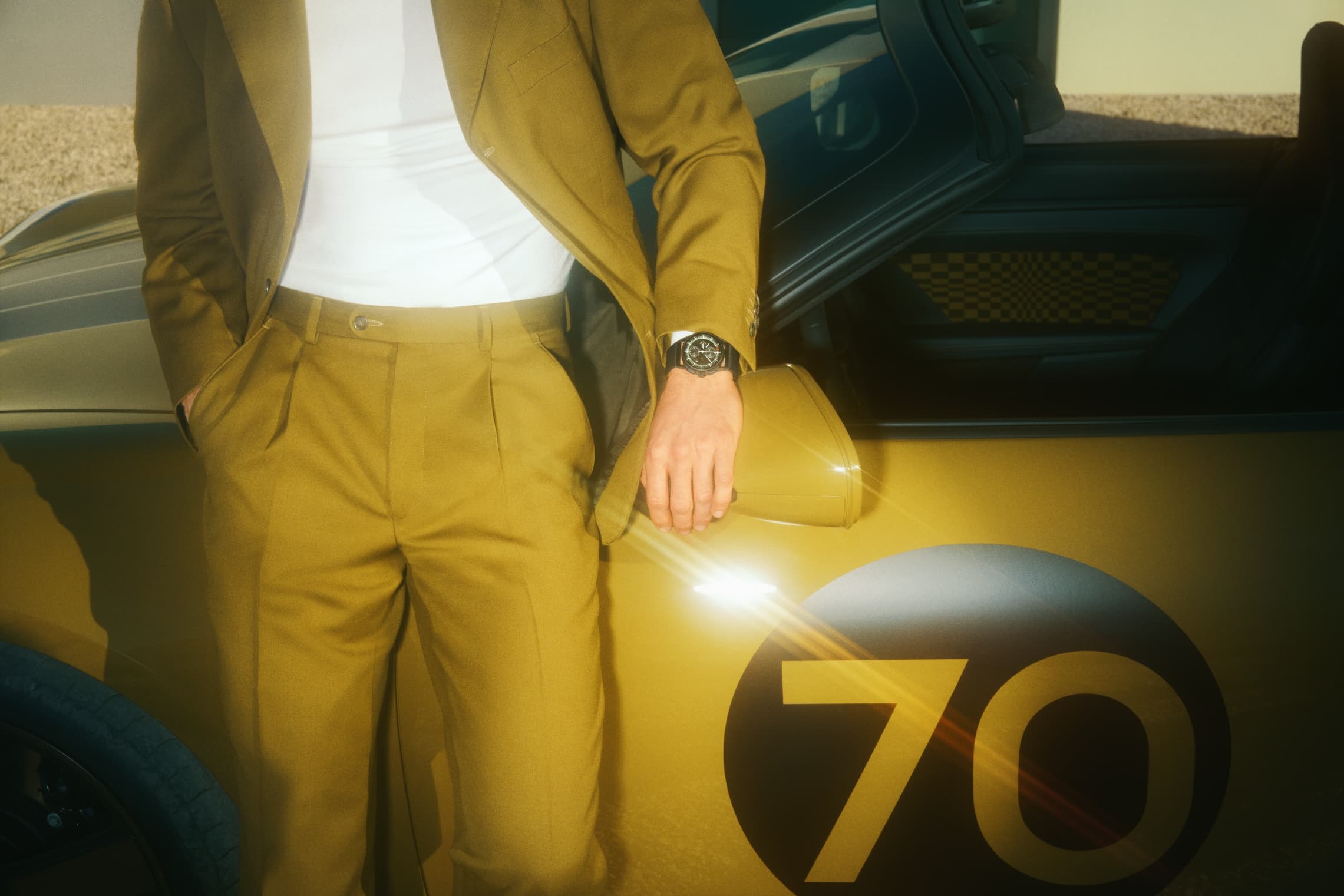 Man in olive-colored suit leaning against a Porsche, wearing the Chronograph 911 Spirit 70 on his wrist