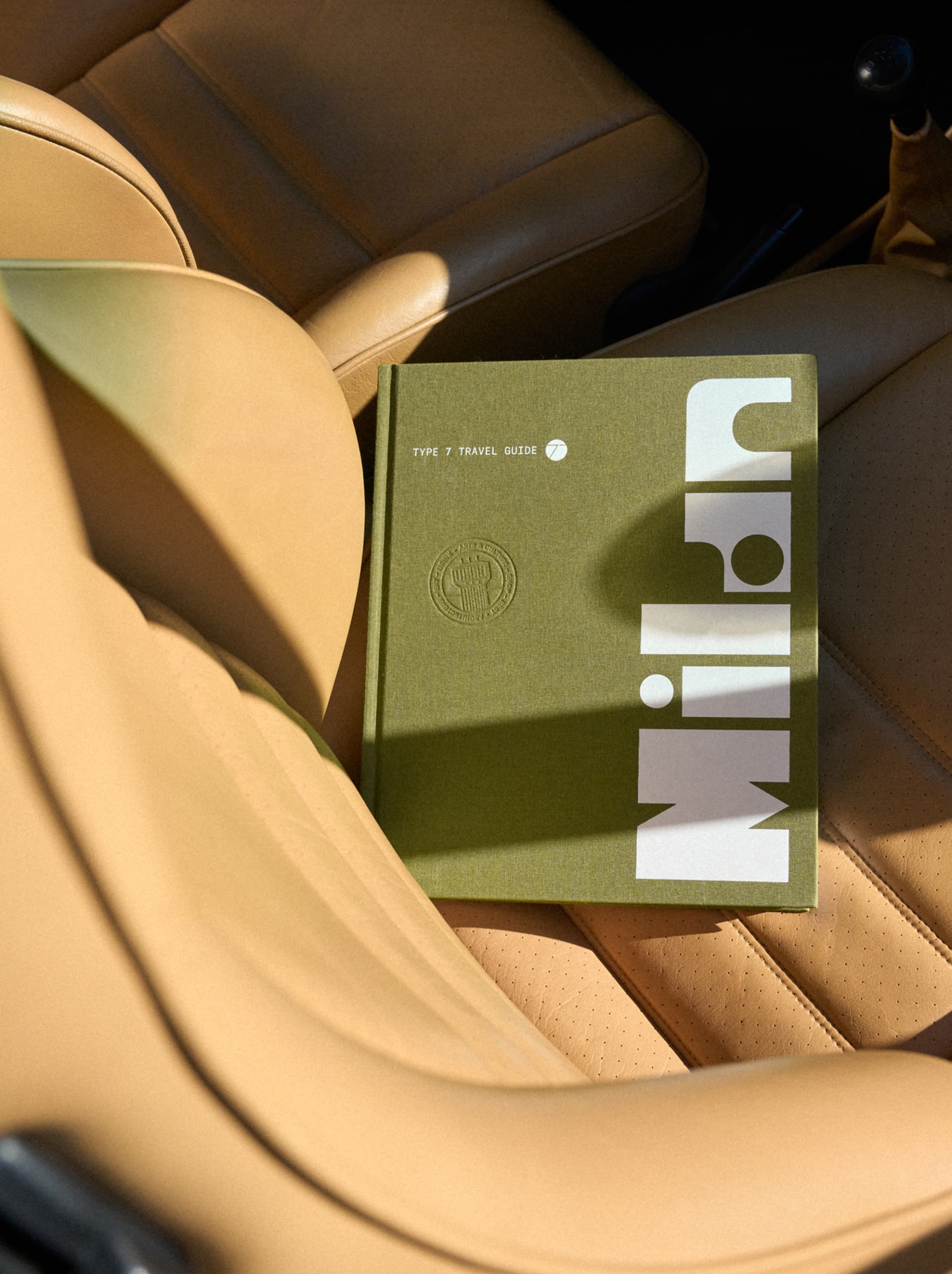 Groen hardcover-reisboek “Type 7 Travel Guide” op een beige lederen autostoel in het zonlicht.
