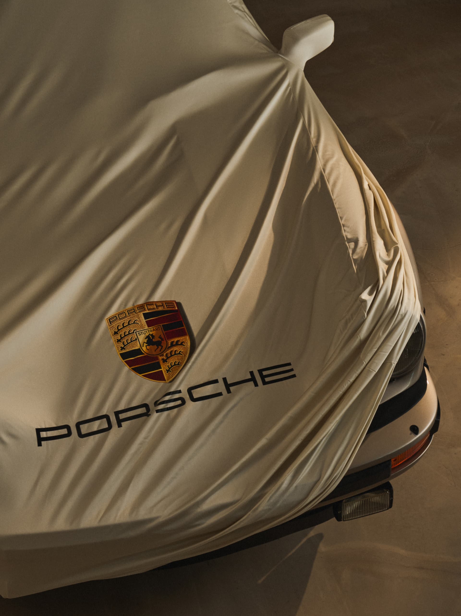 Auto mit beigem Stoff bedeckt, zeigt Porsche-Logo und Text, teilweise die Front des Fahrzeugs enthüllend.