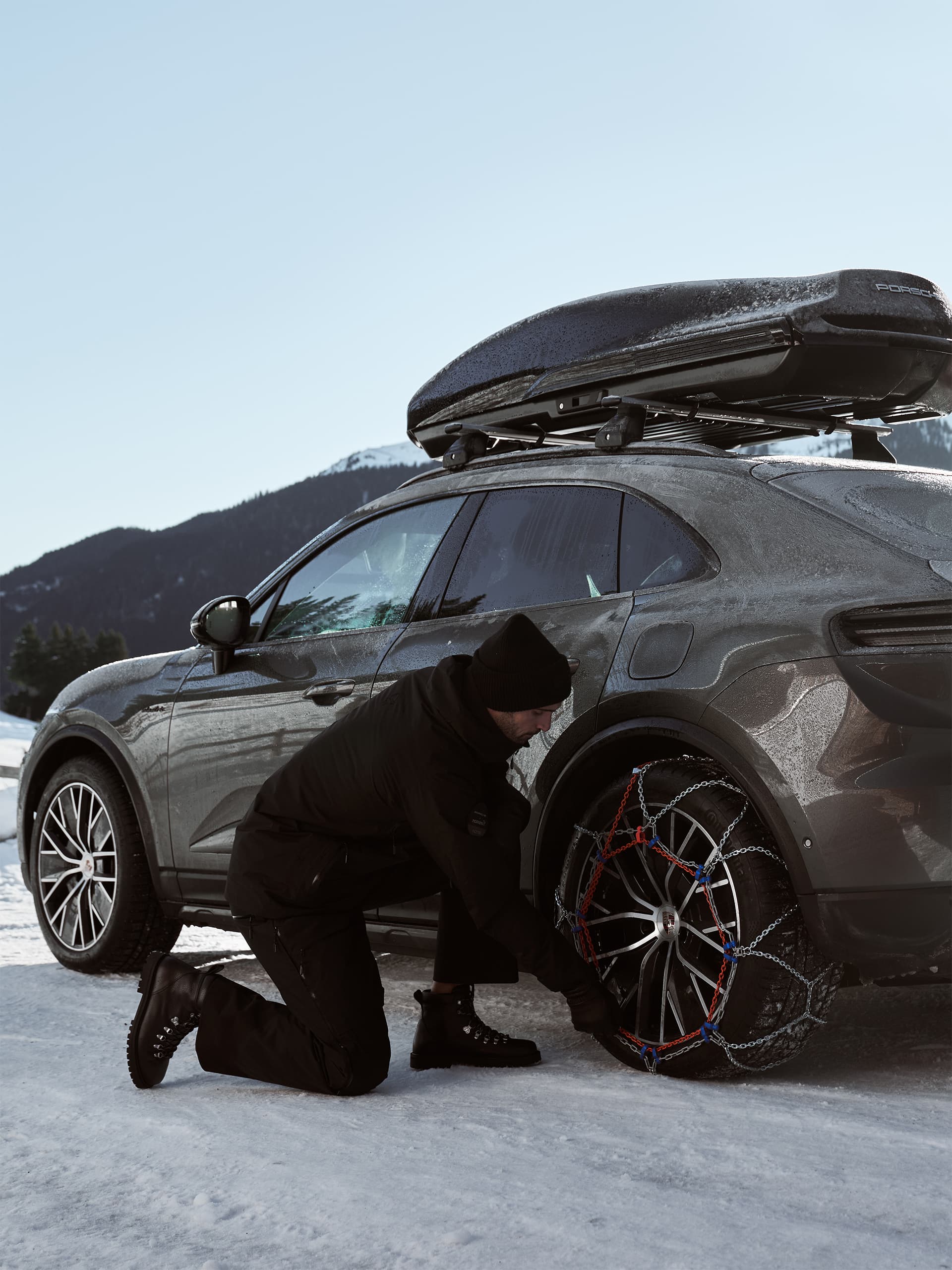 Person fitting snow chains on a gray Porsche with roof box, parked on snowy terrain, mountains in the background.