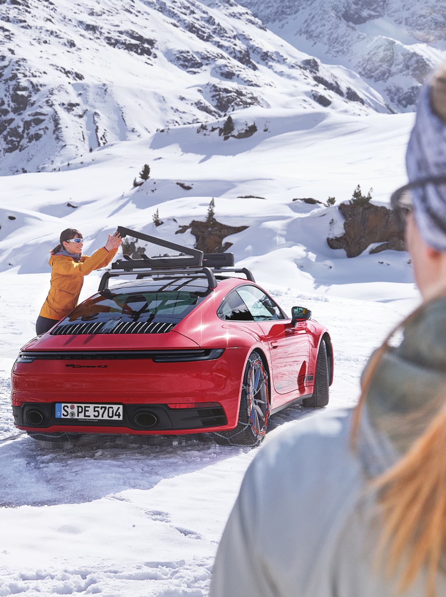 Roter Porsche 911 mit Schneeketten, Person in orangefarbener Jacke passt den Dachgepäckträger an, verschneite Bergkulisse.