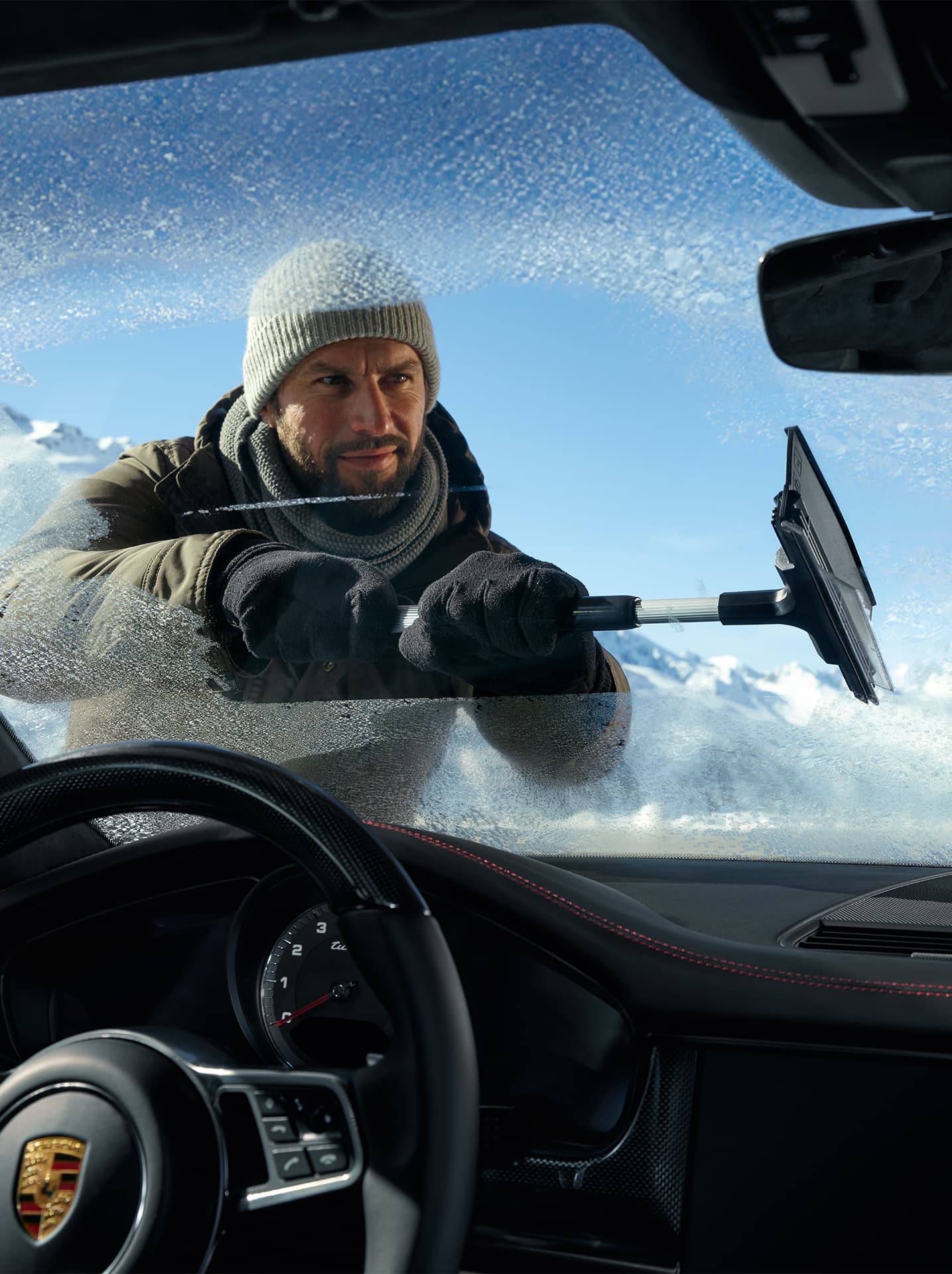 Person fitting snow chains on a gray Porsche with roof box, parked on snowy terrain, mountains in the background.