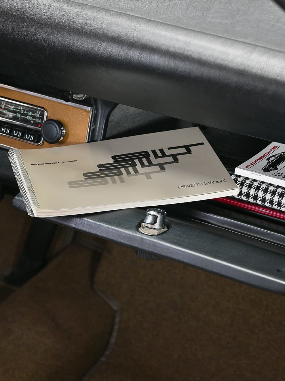 Porsche-Bedienungsanleitung im Handschuhfach, neben einem Buch im Hahnentrittmuster, mit einem nostalgischen Radio im Blick.