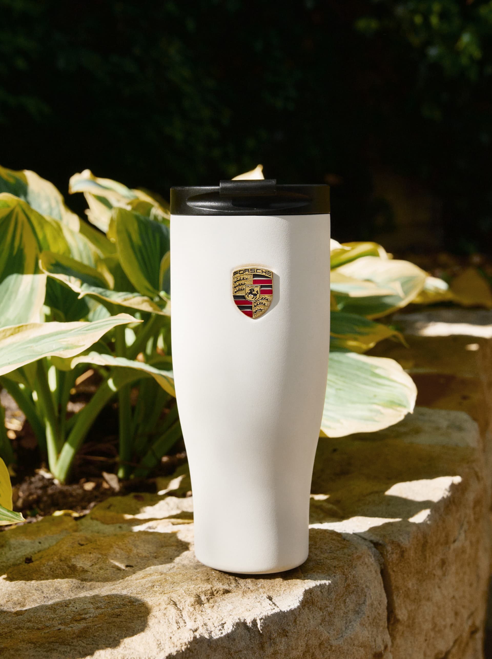 Tazza termica Porsche bianca con coperchio nero e logo con stemma, appoggiata su una pietra davanti a foglie verdi.