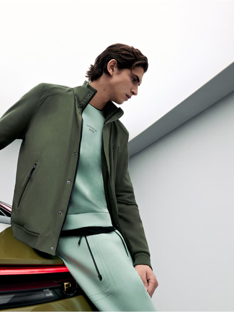 Man in groene jas en mintgroene sweatshirt leunend op een auto, tegen een minimalistische witte en grijze achtergrond.