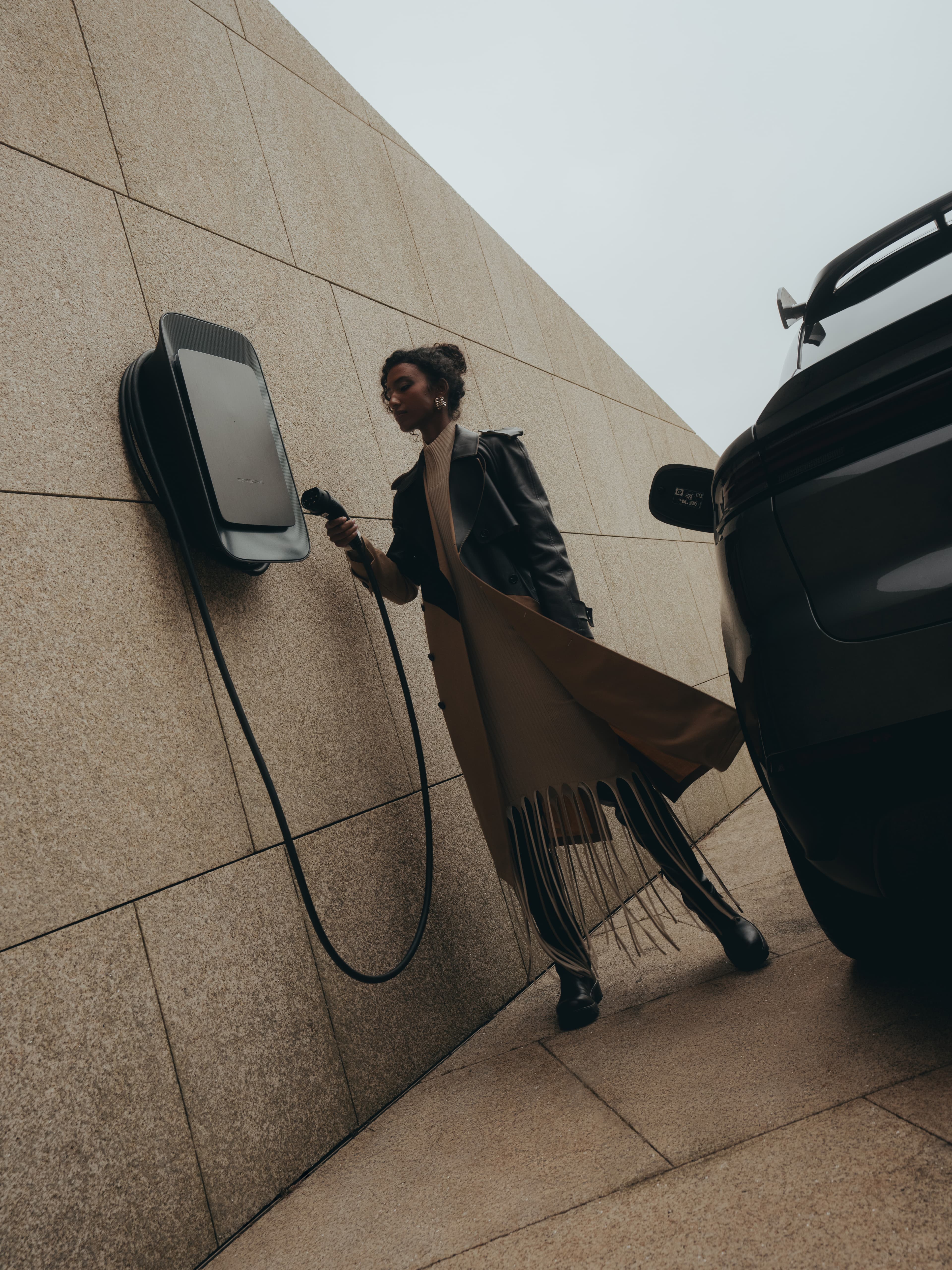 Person in a long coat charging an electric car at a wall-mounted charger on a stone-paved area.