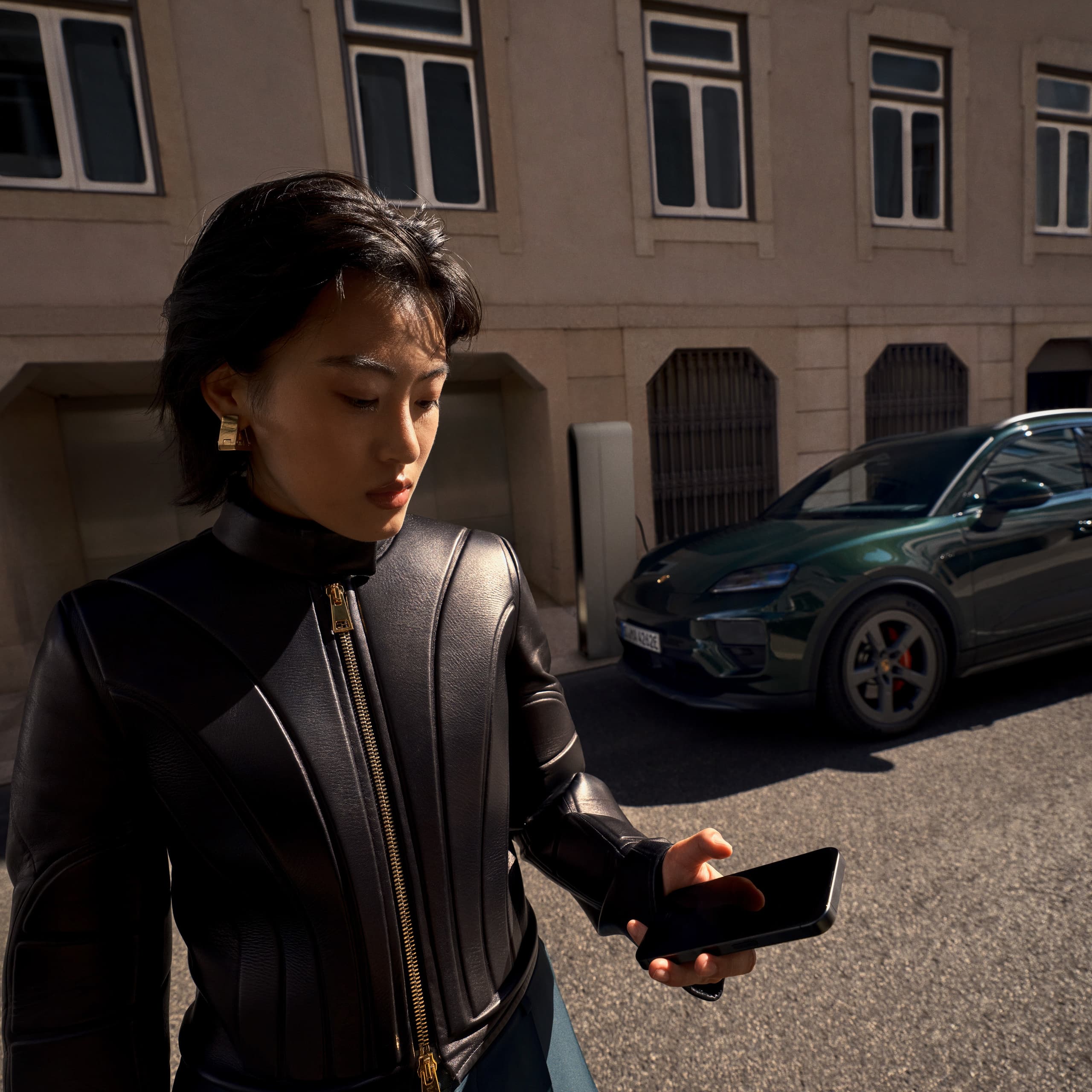 Woman standing in the street holding her mobile phone, a building and a dark green Porsche Macan can be seen in the background