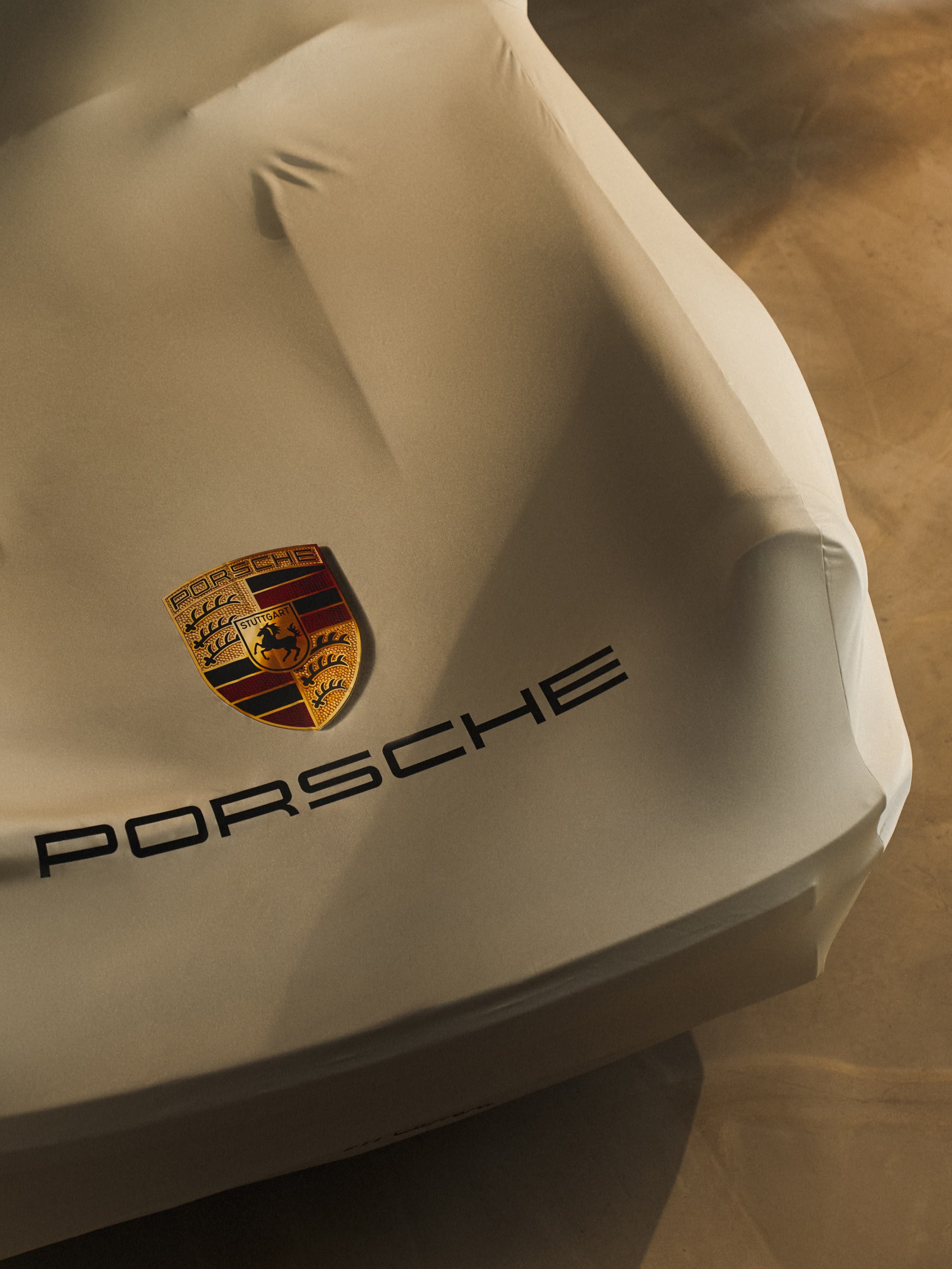 Car covered with beige fabric, displaying Porsche logo and text, on a dimly lit surface.