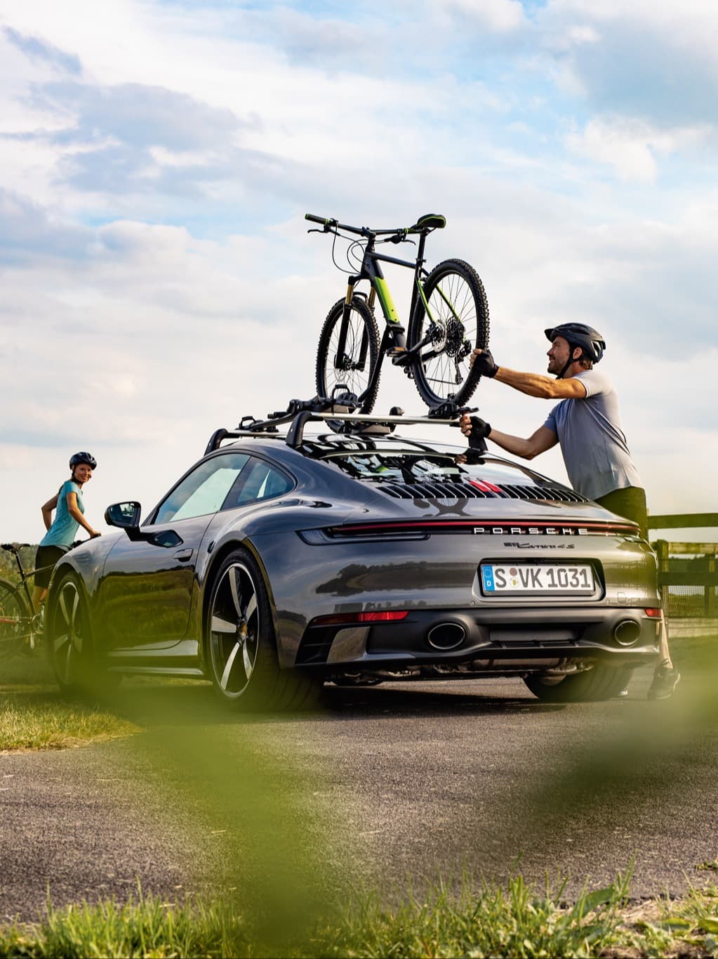 Man secures bike on gray Porsche 911 roof rack, another cyclist nearby, under a cloudy sky.