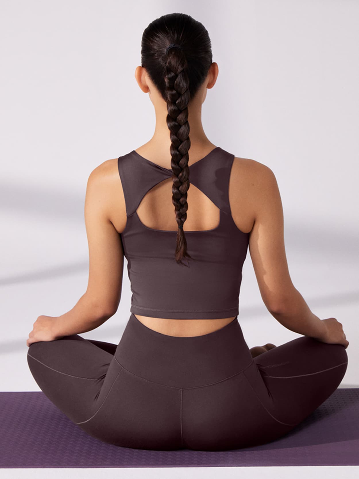 Frau in dunkler Sportbekleidung sitzt im Schneidersitz auf einer lila Yogamatte, mit langem Zopf, von hinten gesehen.