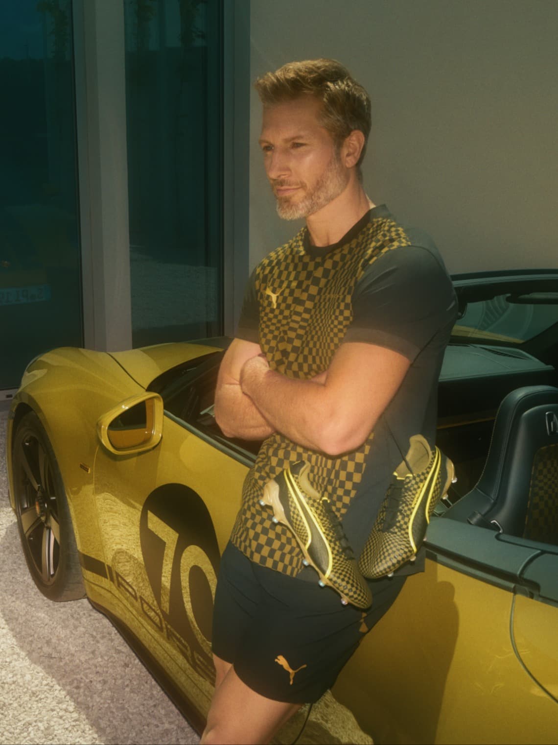 Man in black-and-yellow sports outfit wearing soccer cleats, leaning against a yellow Porsche convertible with arms crossed.
