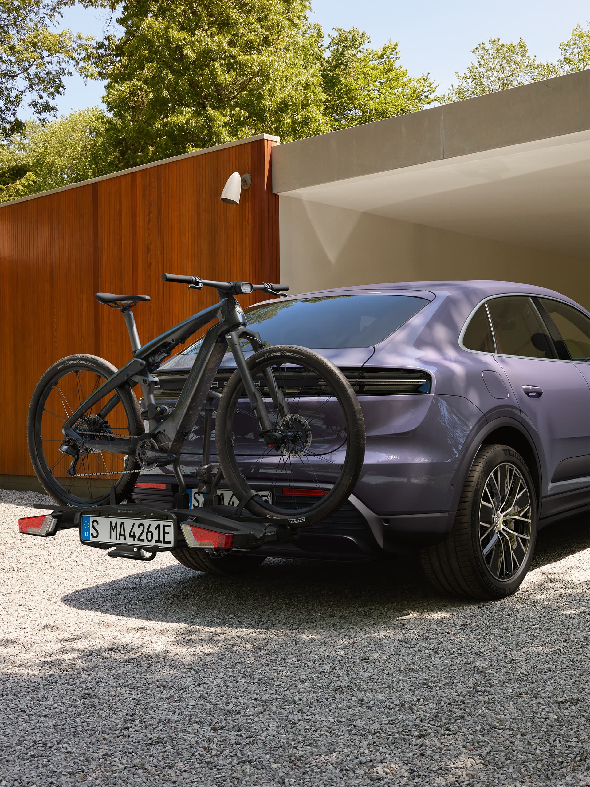 Lila Porsche SUV hátsó kerékpártartóval és rá rögzített fekete elektromos kerékpárral, modern garázs előtt parkolva, zöld növényzettel körülvéve.