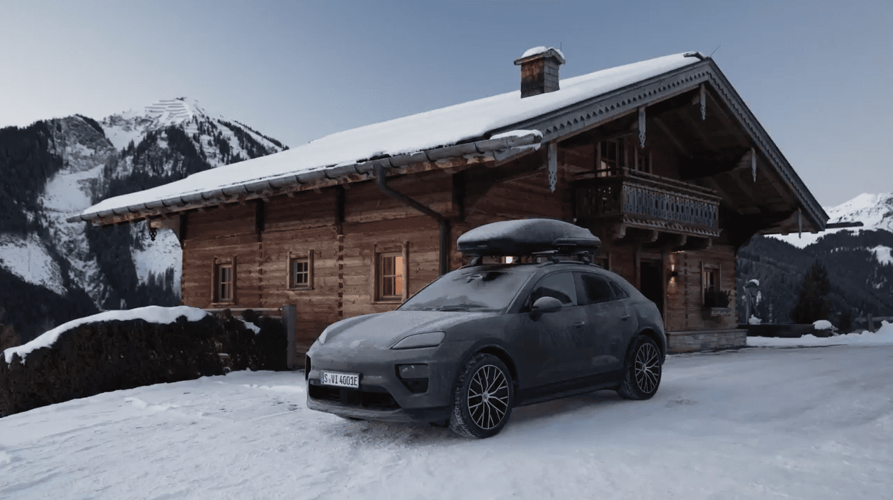 Porsche SUV with roof box parked by snowy wooden chalet, mountain backdrop